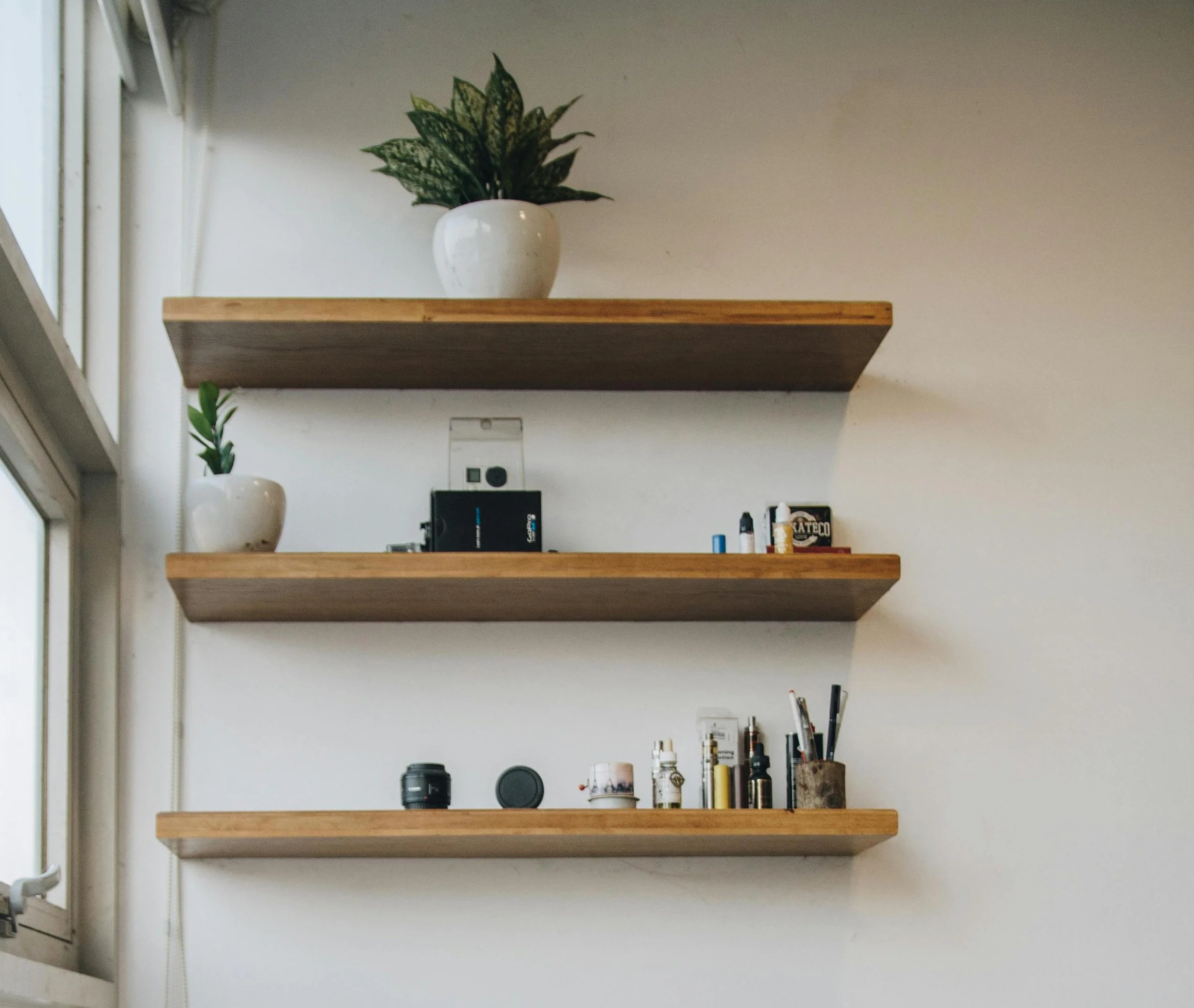 Three wooden shelves mounted on a white wall with potted plants on the top and middle shelves, and various small items including bottles, a camera lens, and containers on the bottom shelf.