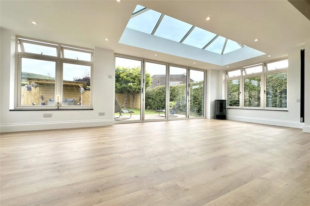 Empty living room with large windows, glass doors leading outside, a skylight, wooden flooring, and minimal decor.