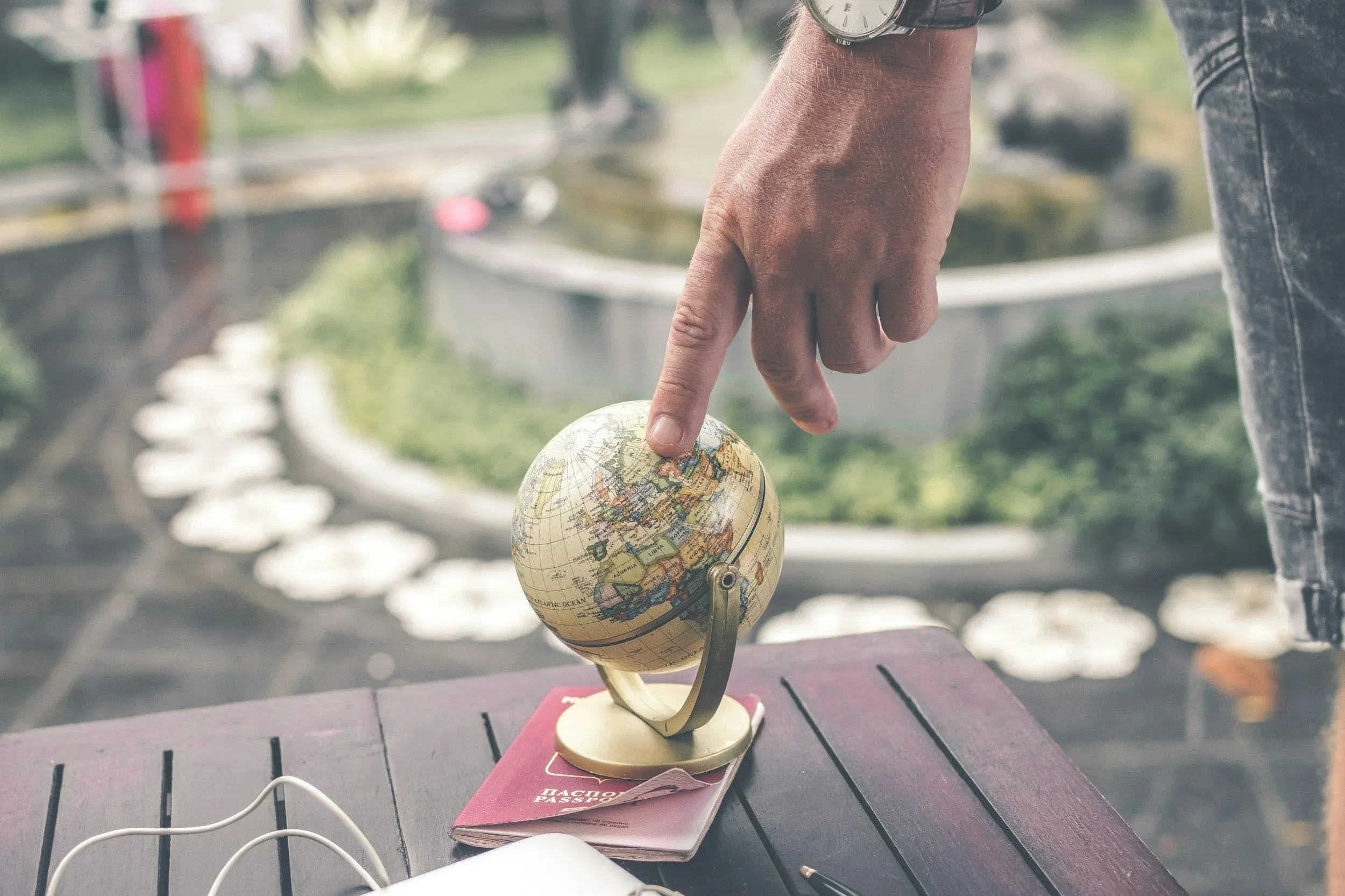 A person's hand is pointing towards a globe on a table. The globe is placed on top of a red passport and a white object, with earphones visible nearby. The scene appears to be near a window with an outdoor area in the background.