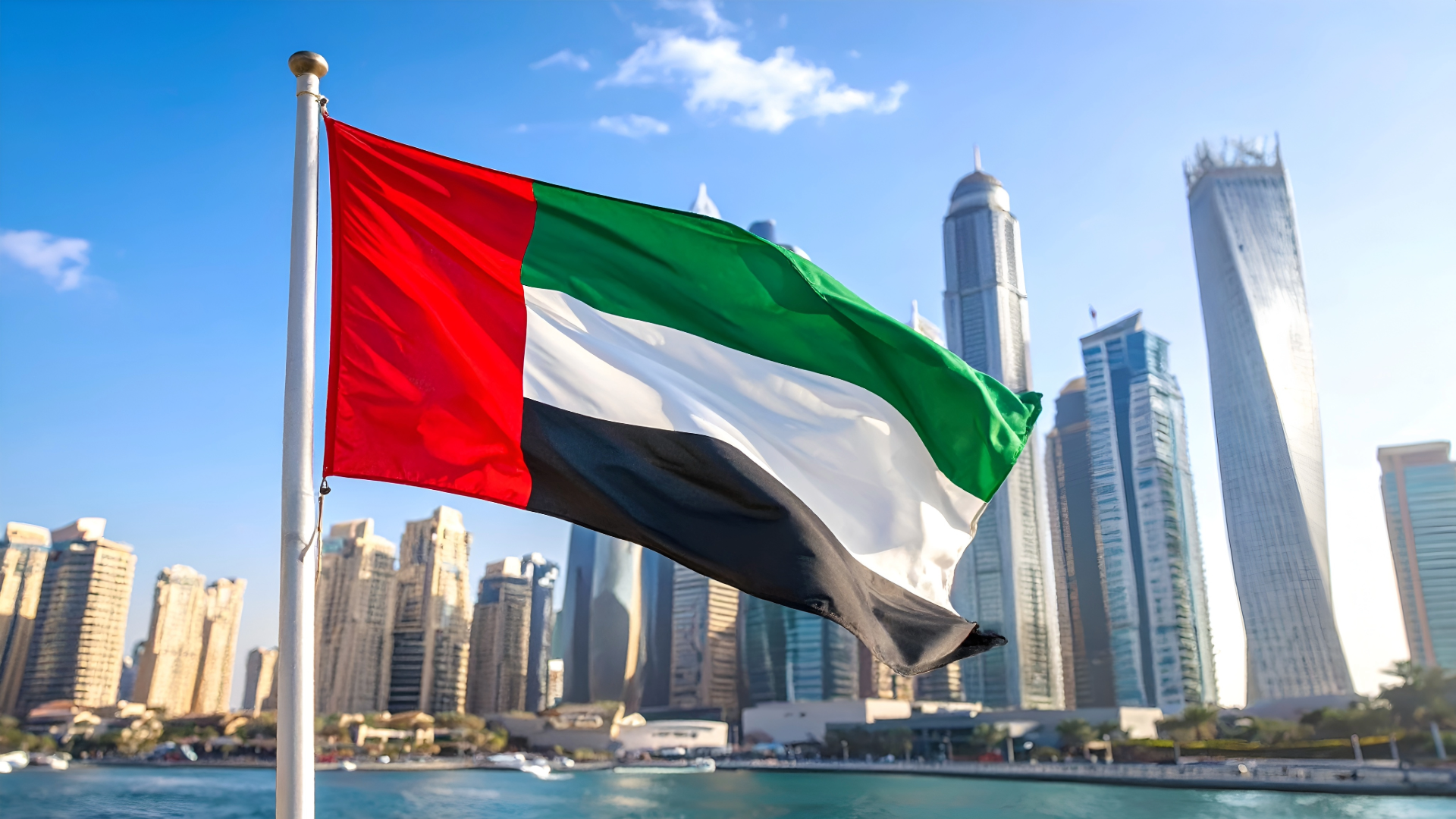 UAE flag waving in front of Dubai skyline with tall skyscrapers and water in the foreground.