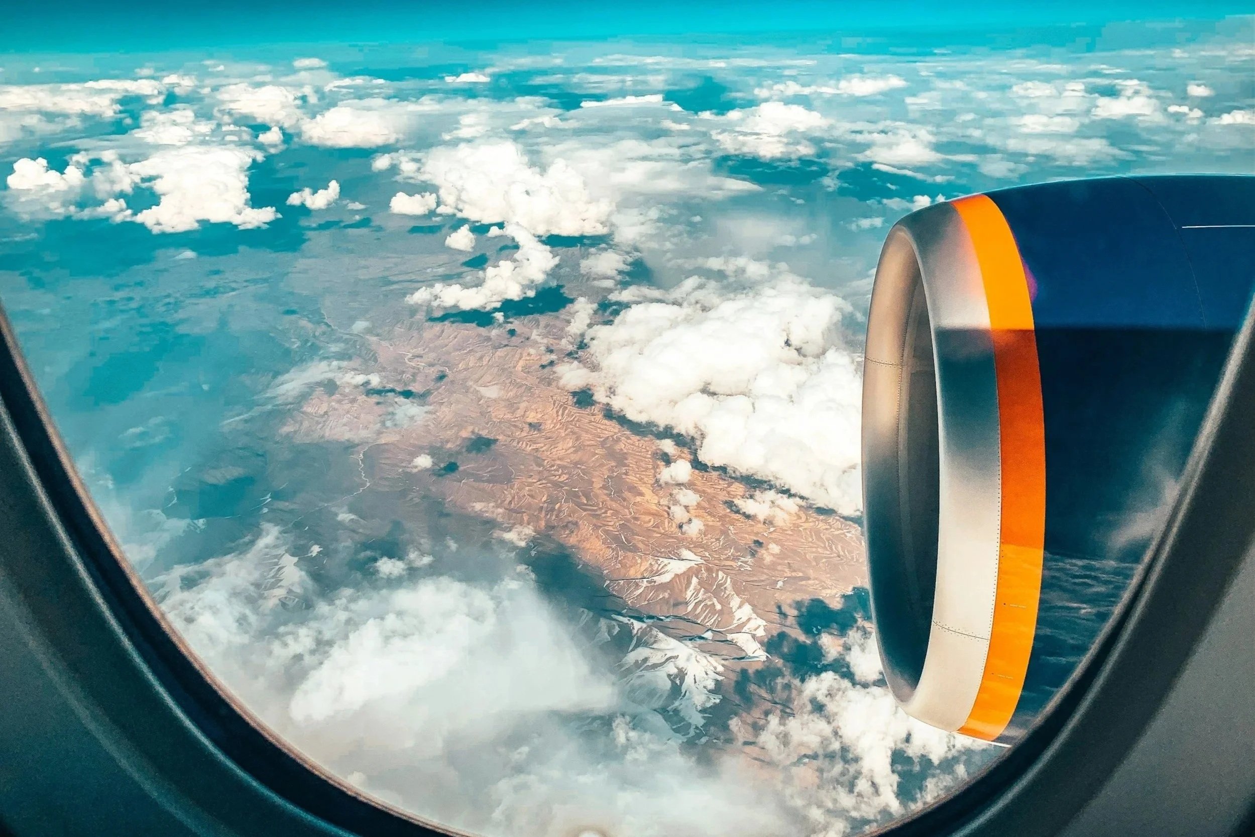 View of a landscape with clouds and mountains from an airplane window, showing part of the jet engine with an orange stripe.