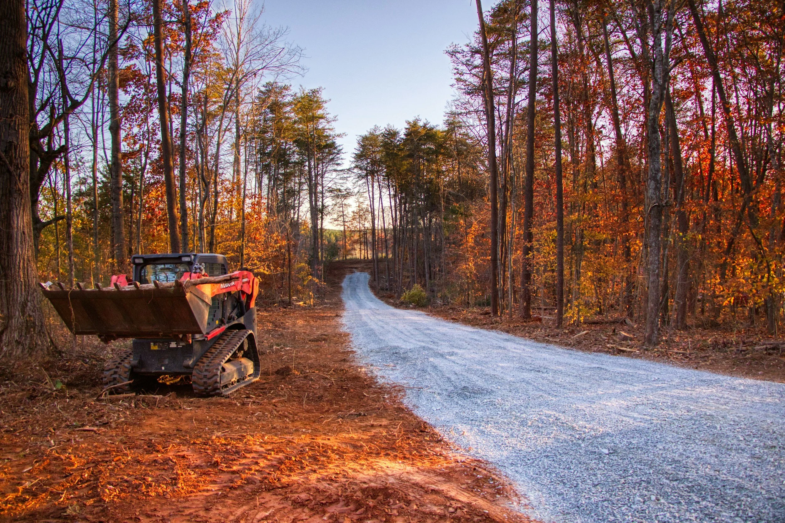 A gravel road winding through a forest with trees displaying autumn colors. A small tracked construction vehicle is parked on the side of the road.