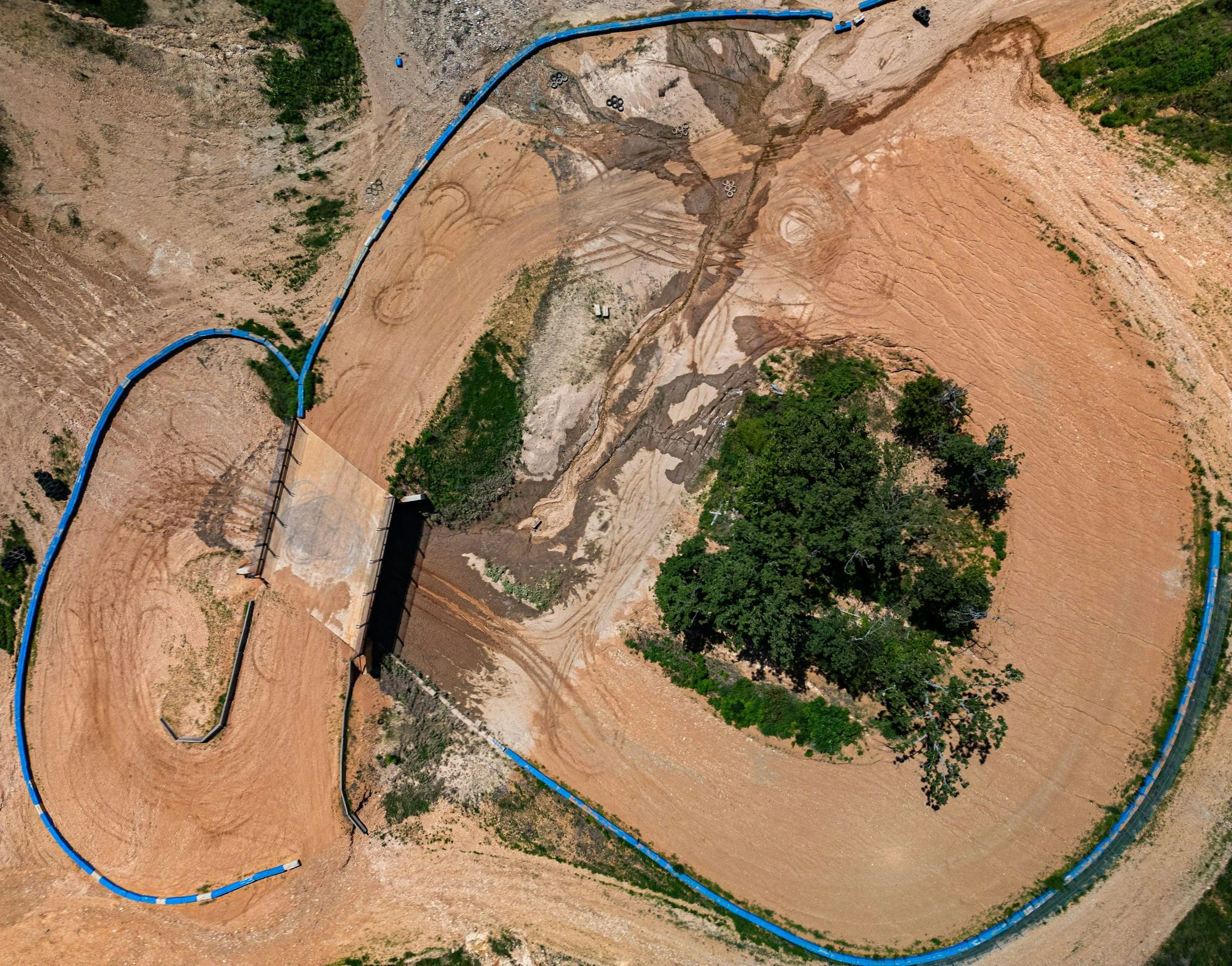 An aerial view of a construction site with a dirt track, a building foundation, and scattered trees, surrounded by a blue barrier.