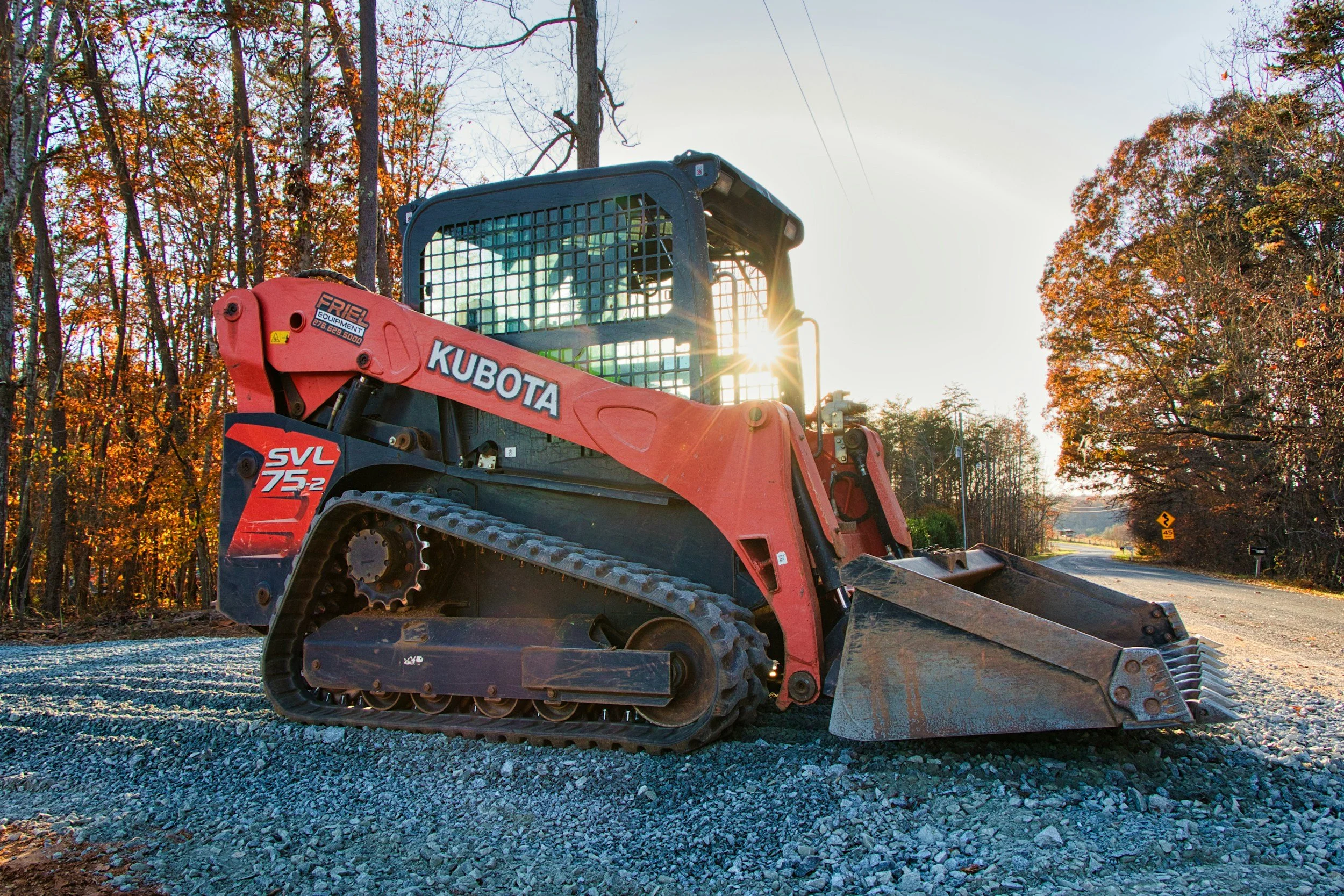 A Kubota SVL75-2 compact track loader parked on a gravel surface with autumn trees and a road in the background, with sunlight shining through the trees.