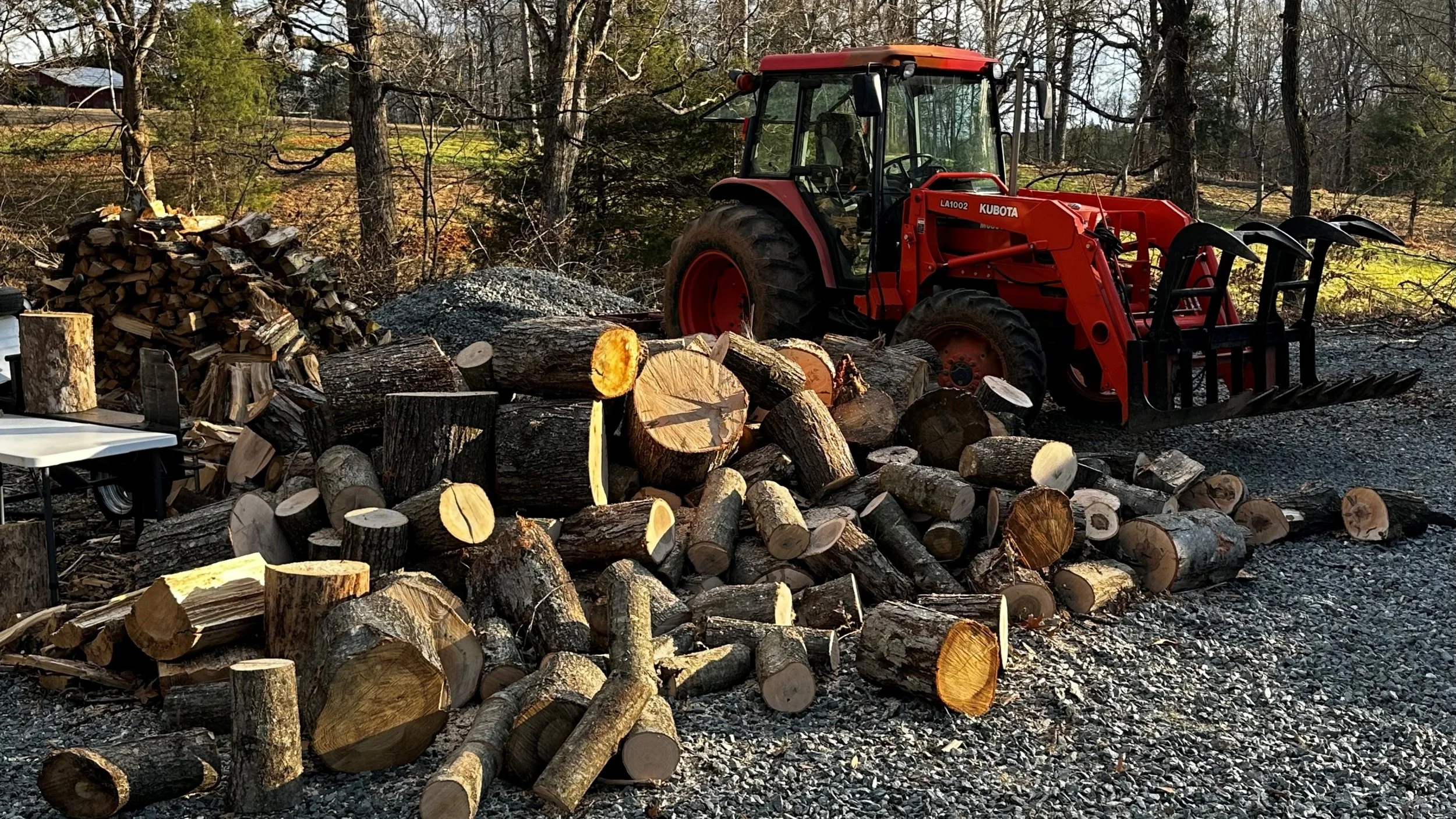 Firewood and logs stacked outdoors with a red tractor nearby.