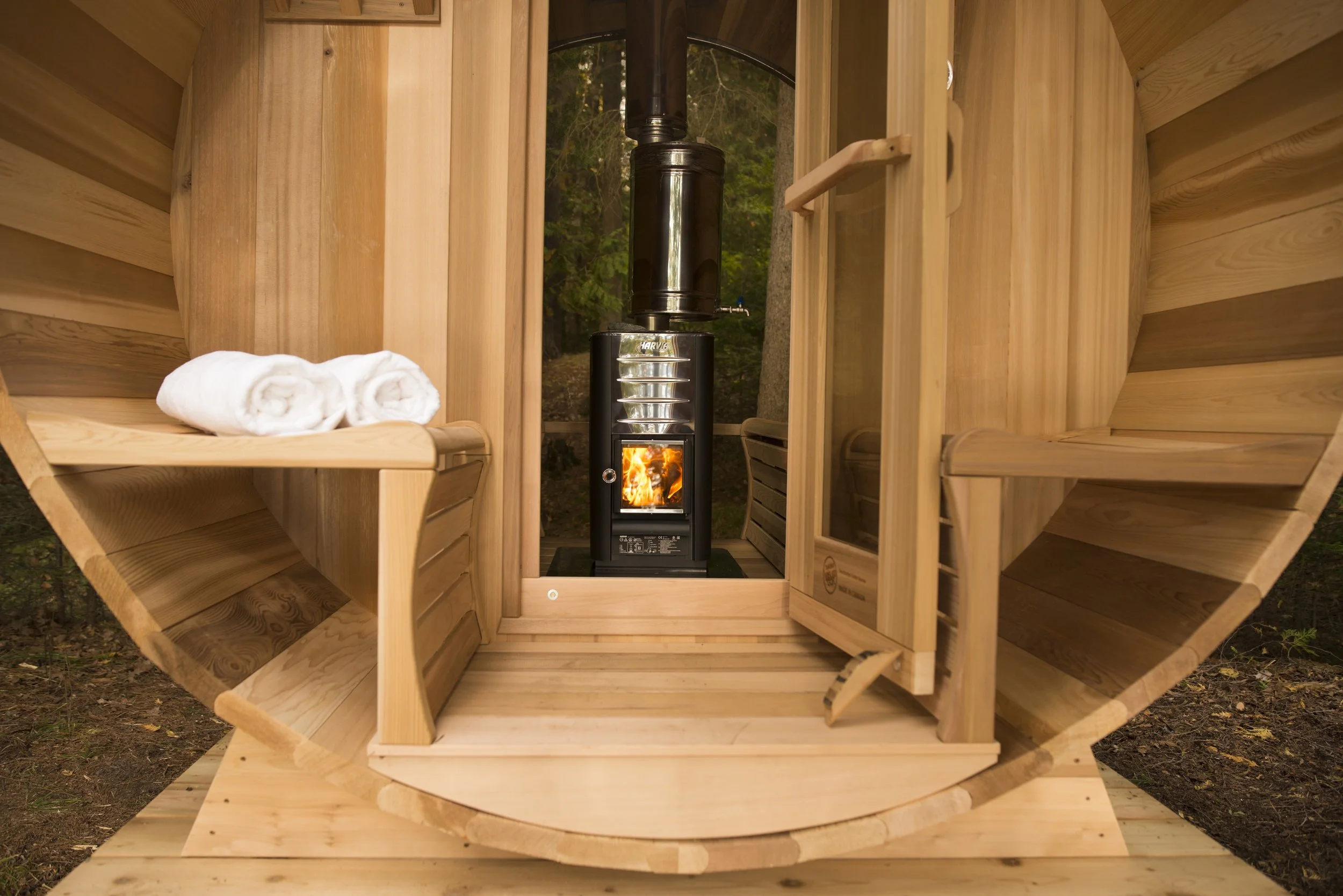 Inside a small wooden sauna with a wood stove burning fire, and two rolled towels on a bench.