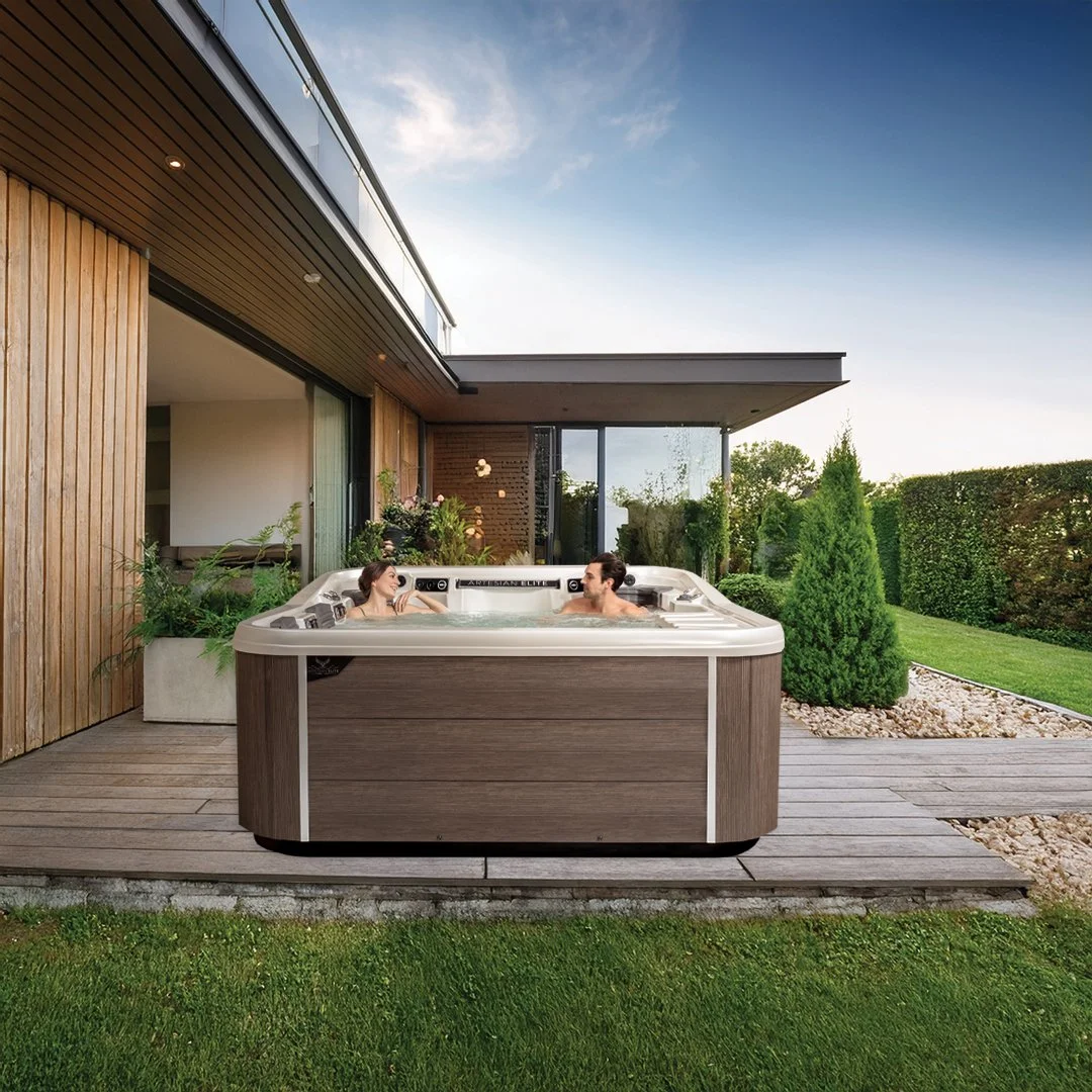 Two people relaxing in a hot tub on a wooden deck next to a modern house with large glass windows and a backyard garden with shrubs and trees.