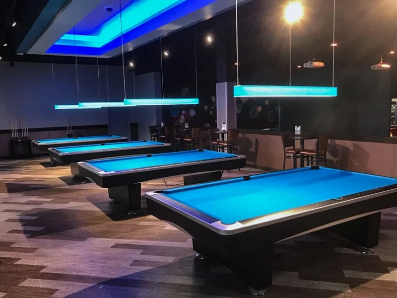 A modern indoor pool hall with four pool tables illuminated by blue hanging lights. The room has dark walls, a patterned wooden floor, and seating along the back wall.