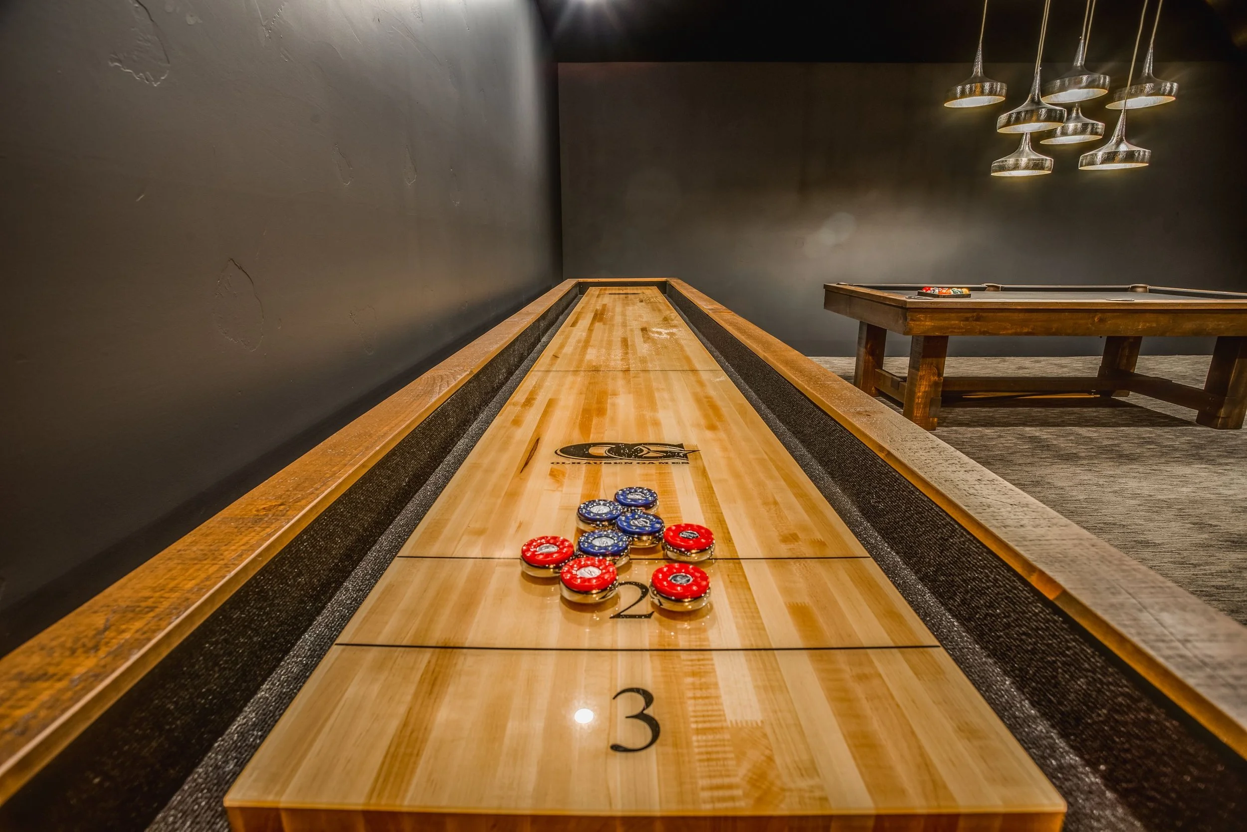 A wooden shuffleboard table with a set of shuffleboard pucks in red and blue, numbered 1, 2, and 3. The table is in a dimly lit room with dark-colored walls and a set of modern pendant lights hanging from the ceiling.
