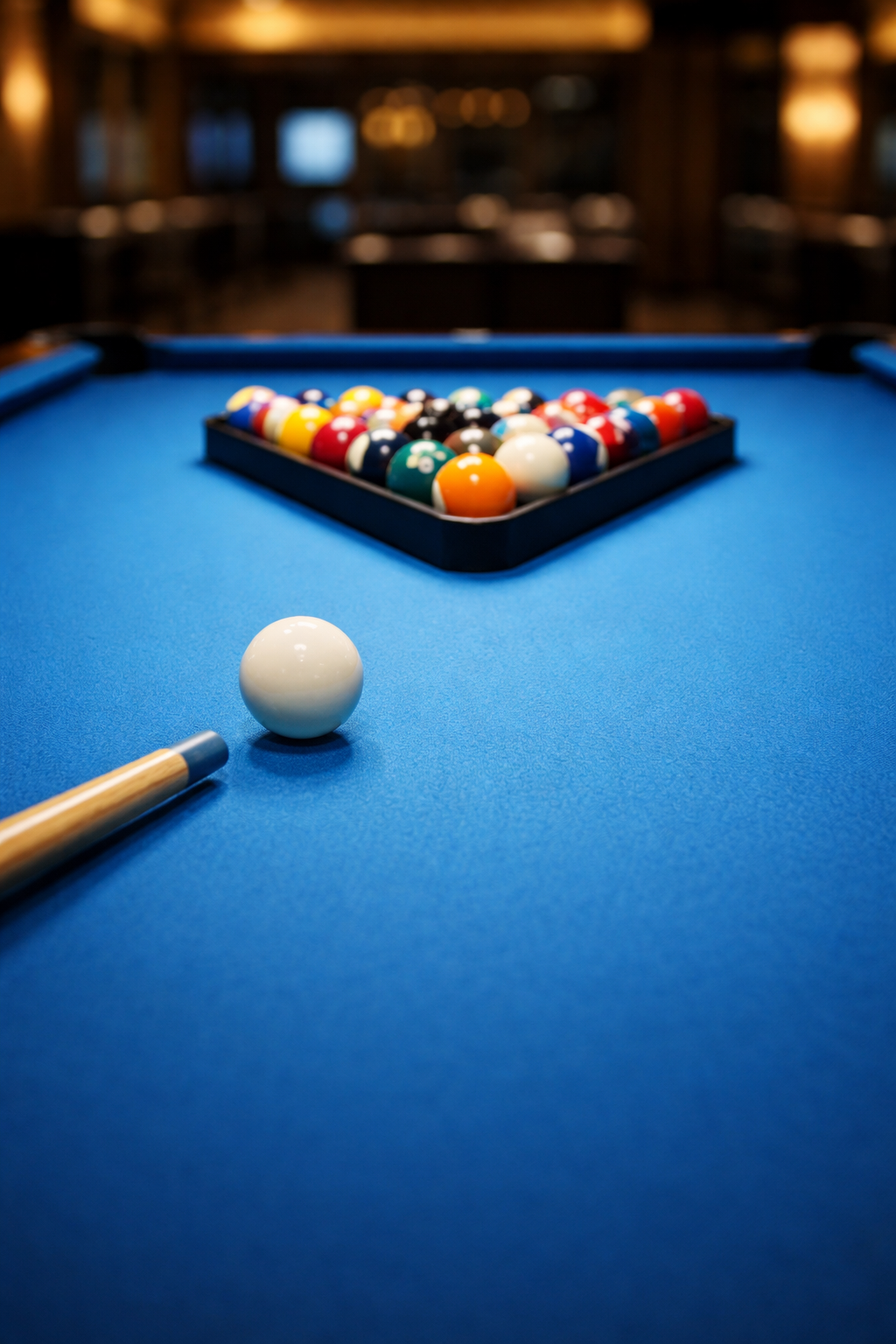 A pool table with a rack of pool balls arranged for a game, a cue stick, and the cue ball in position on the blue felt surface.