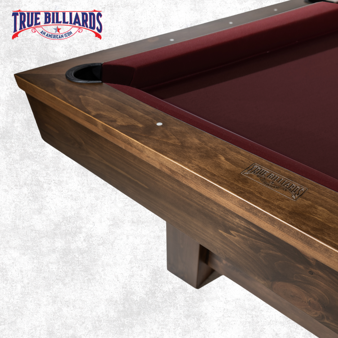 Close-up of a pool table with a wooden frame, red felt surface, and a corner pocket. The table has a carved "True Billiards" logo on the wood.