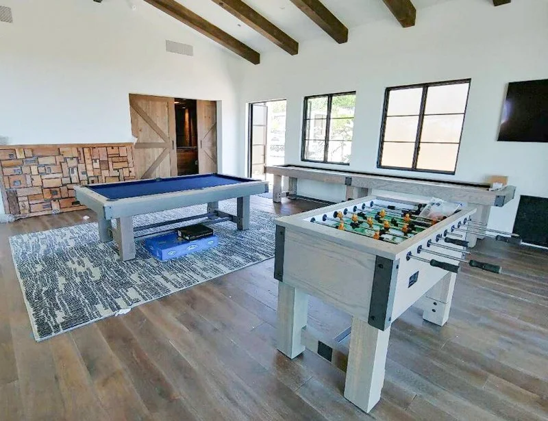 Game room with pool table, foosball table, and large windows in a sunny space with wooden beams and flooring.