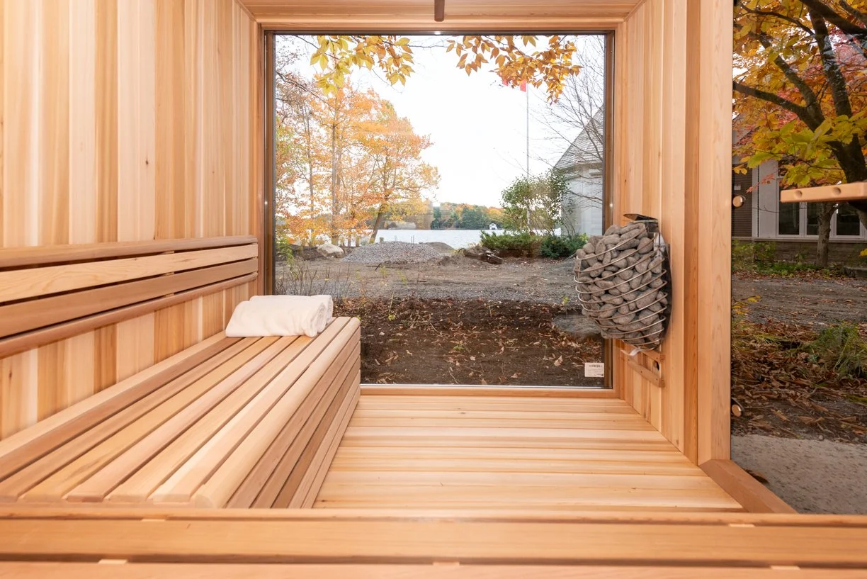 Sauna with wooden interior, large window showing fall trees and outdoor landscape, towel on bench, heated rocks for steam inside sauna.