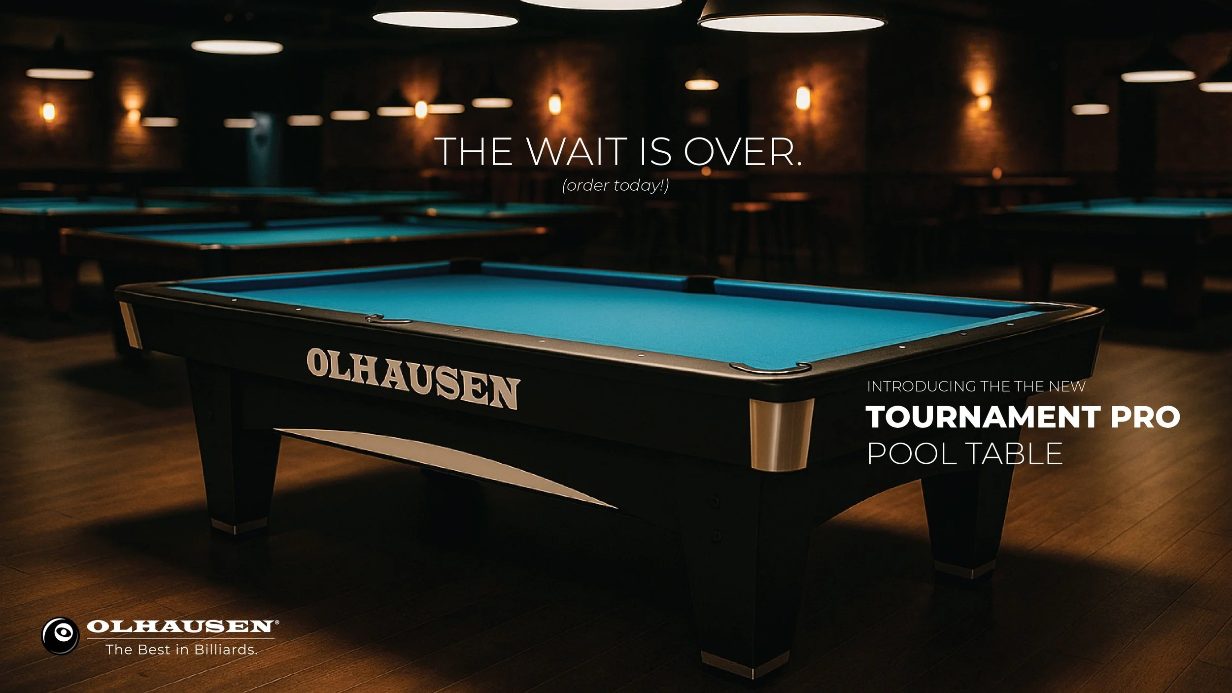 A pool table in a dimly lit room with brick walls and modern lighting. The pool table is branded Olhausen and features a blue felt surface, along with promotional text about a new Tournament Pro Pool Table.