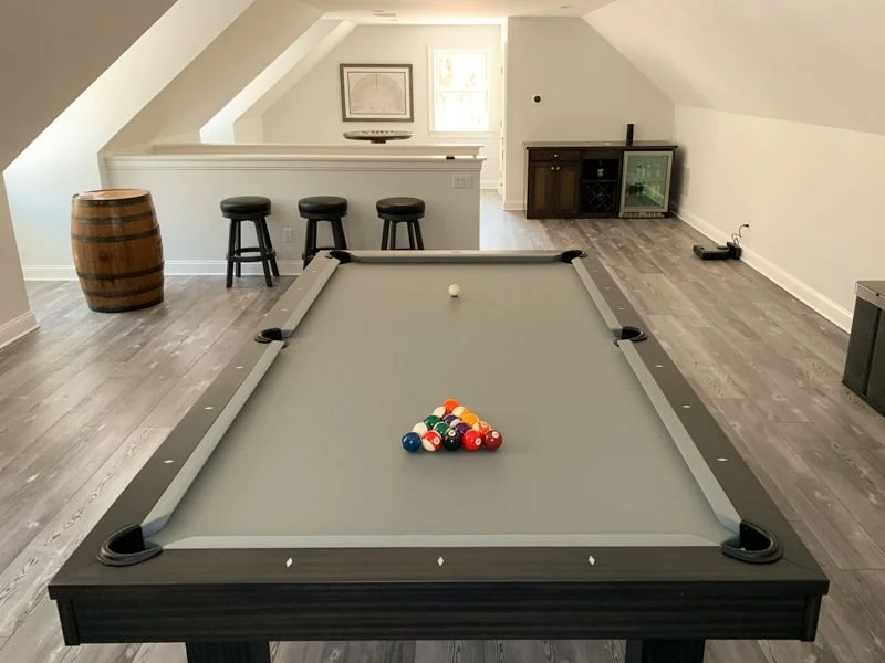 A pool table with balls racked on it in a room with wooden floors, a bar area with barstools, and a small cabinet with a fish tank in the background.