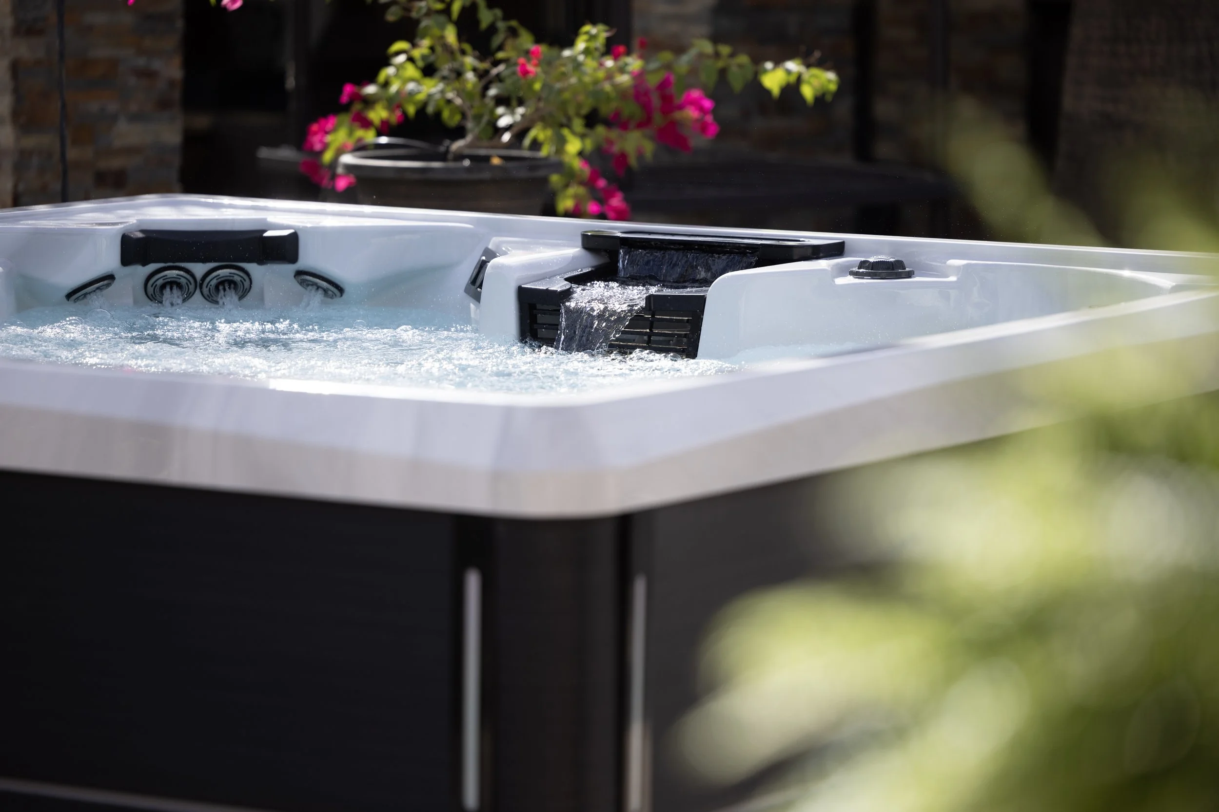 An outdoor hot tub with bubbling water and built-in jets, set against a background of brick walls and a potted flowering plant with pink blossoms.