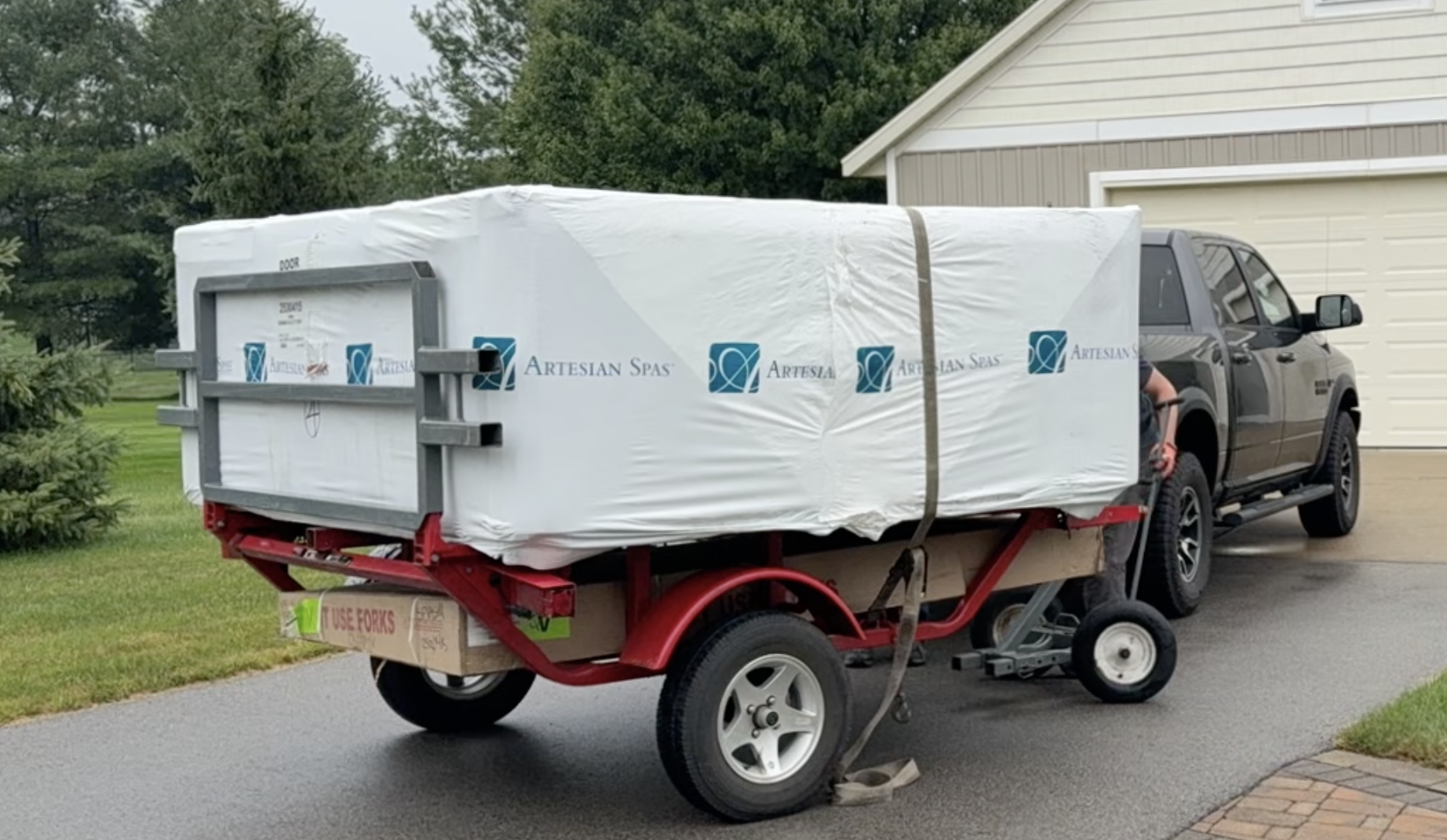 A pickup truck parked on a driveway hauling a trailer with a white wrapped spa labeled 'Artesian Spas.' The trailer has red and black components and a sign that says 'Use Forks.' There is a house with a beige garage door and trees in the background.