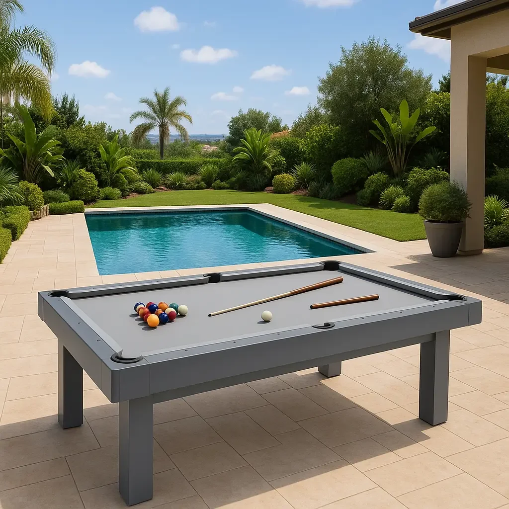 Poolside view with a billiards table in the foreground, pool, and lush green tropical plants and trees under a partly cloudy sky.