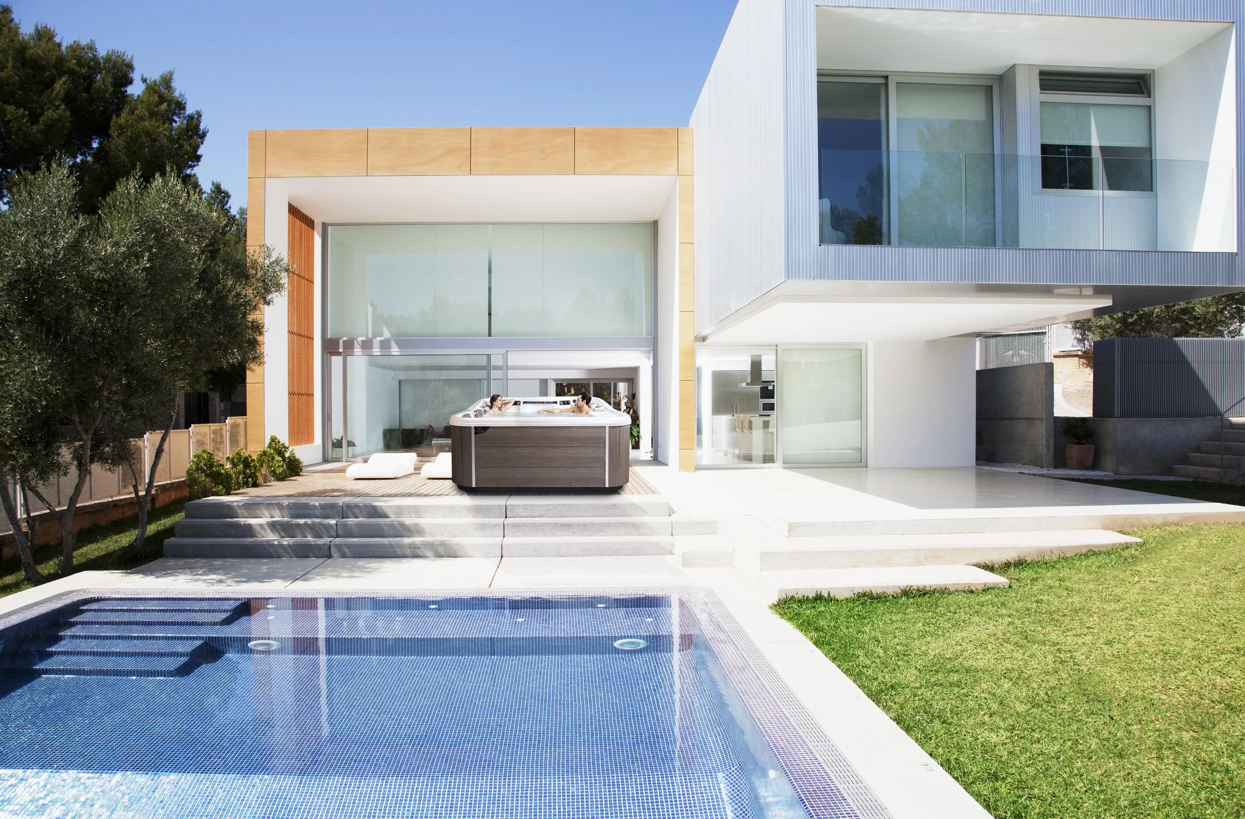 Modern white house with large glass windows, outdoor hot tub on patio, swimming pool in foreground, green lawn, and trees in the background under a clear blue sky.