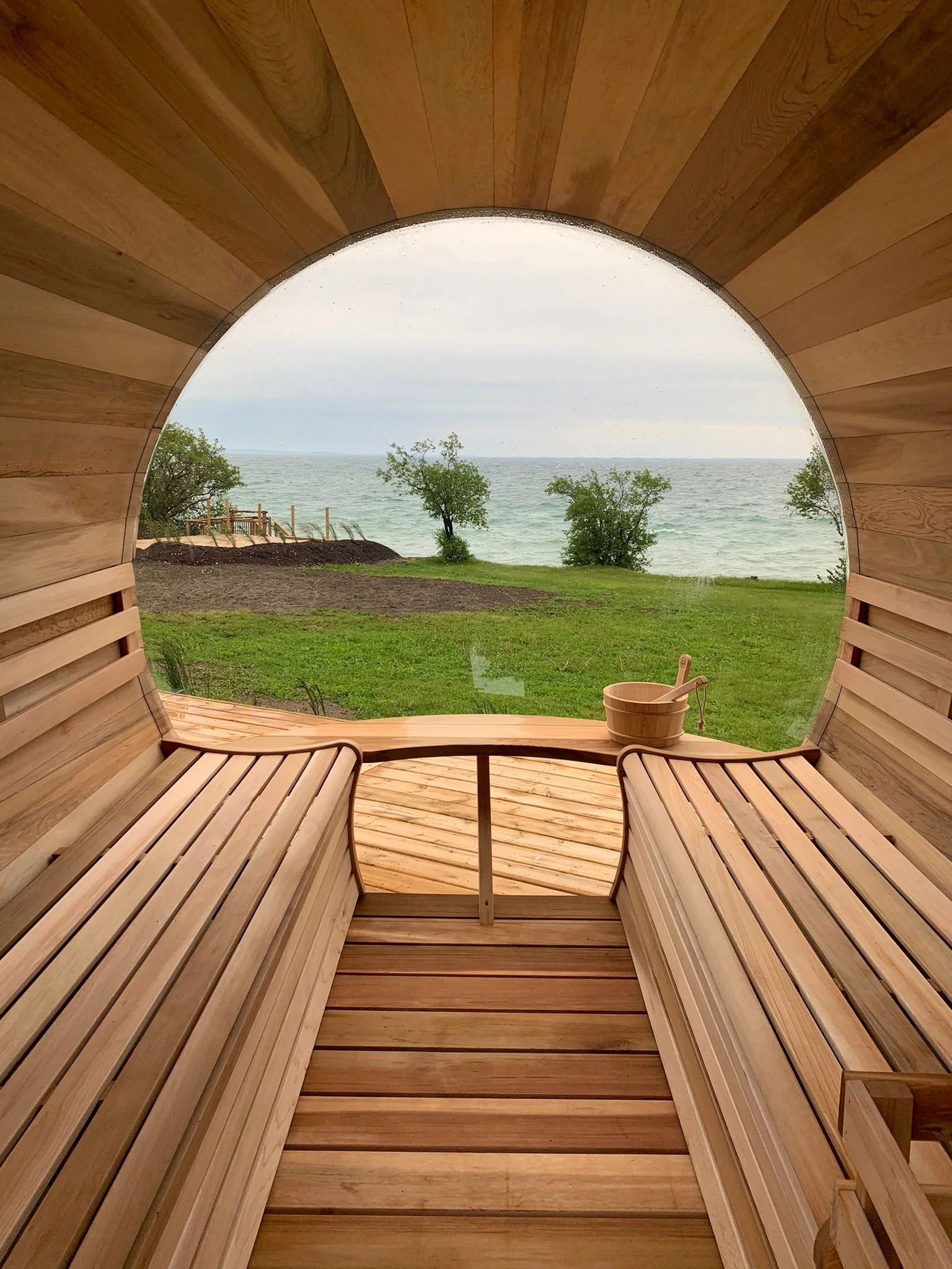 Interior view of a wooden sauna with benches and a view of the outdoors through a circular opening showing trees, water, and the sky.