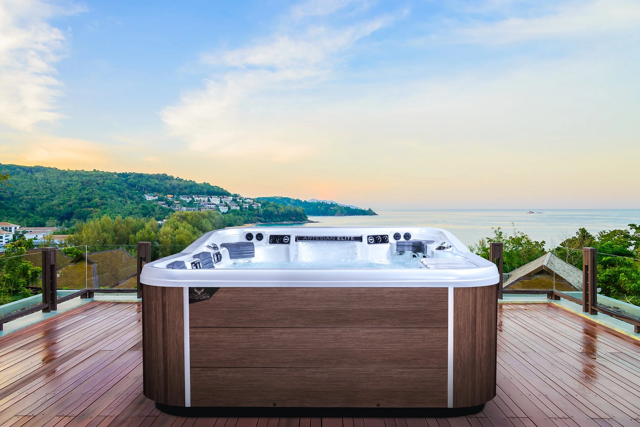 Outdoor hot tub on a wooden deck overlooking a scenic landscape with water, houses, trees, hills, and a partly cloudy sky during sunset.