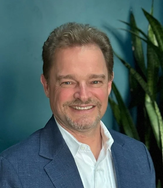 Headshot of Jonathan Ramsden smiling with a blue wall and plant in the background.