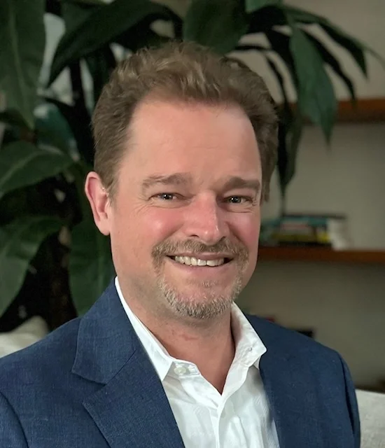 A Headshot or Jonathan Ramsden smiling in front of a plant.