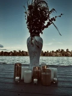 A vase with dried or wilted flowers placed on a wooden surface, with candles surrounding it, by a body of water under a cloudy sky.