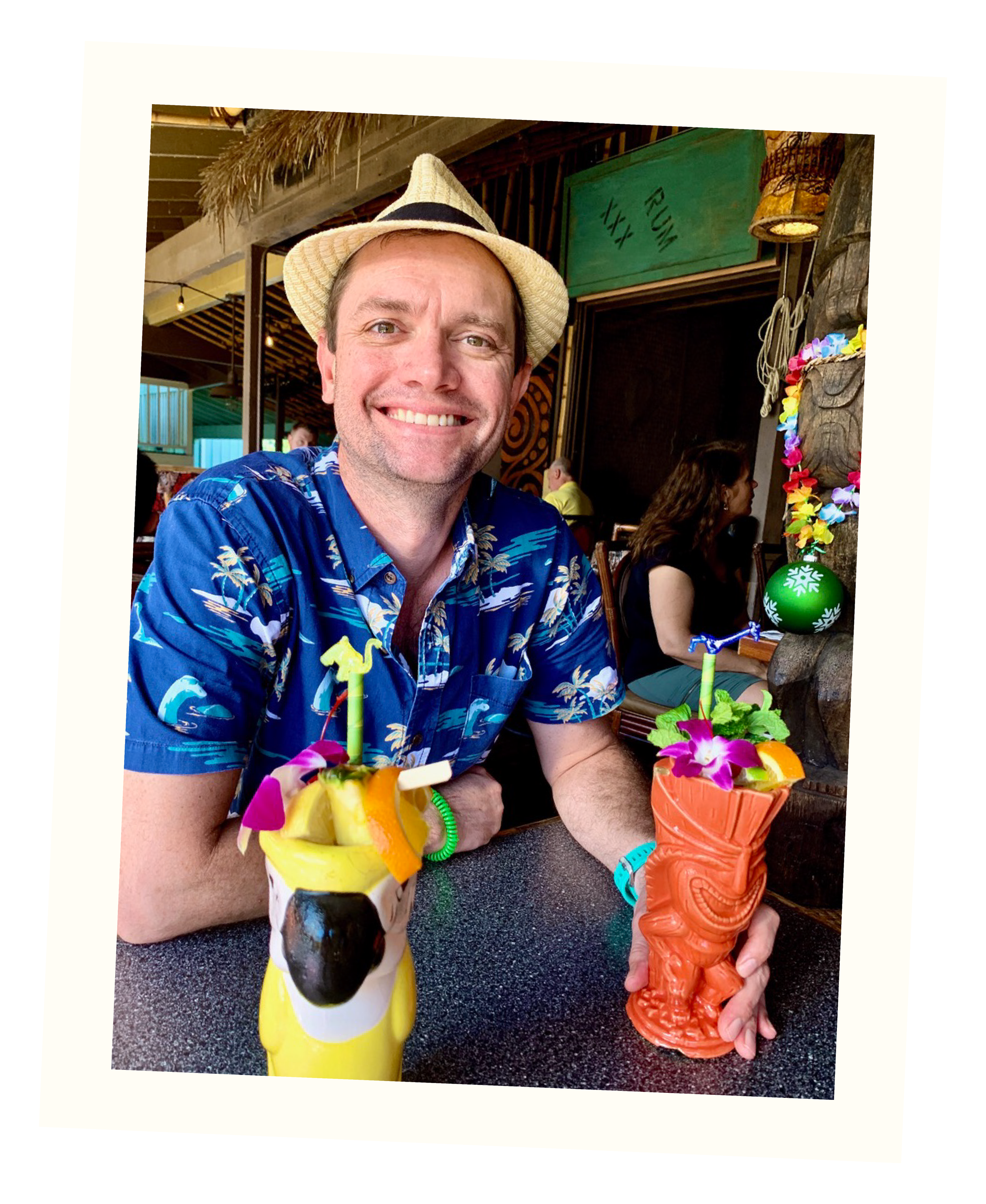A man in a blue Hawaiian shirt and a straw fedora smiling at the camera, sitting at a tropical-themed bar with two colorful tiki drinks garnished with fruit and flowers.