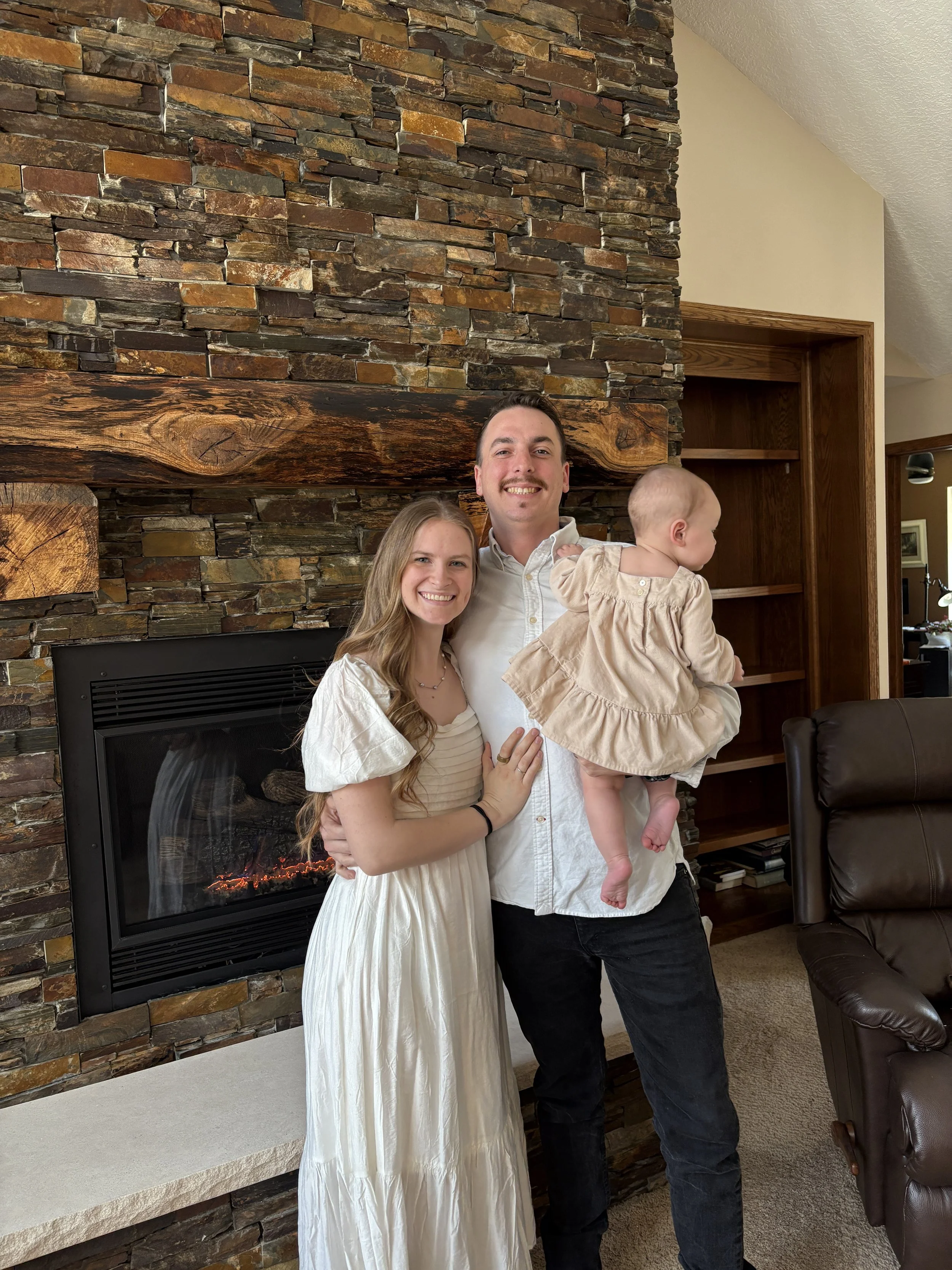 A man holding a young girl on his arm, standing next to a smiling woman in front of a stone fireplace with a wooden mantel in a living room.