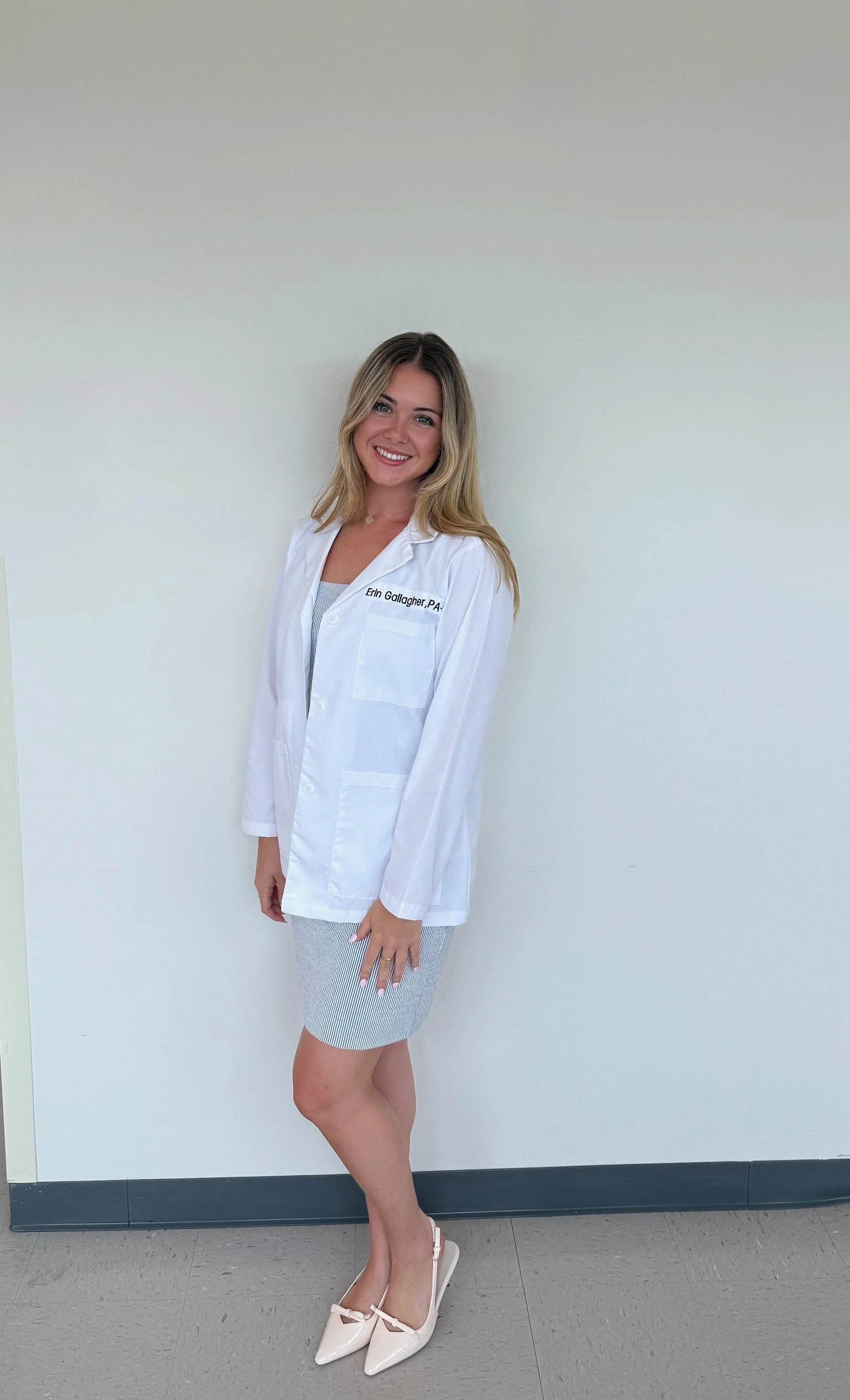 A smiling woman with blonde hair wearing a white lab coat and a name tag standing in front of a plain white wall.