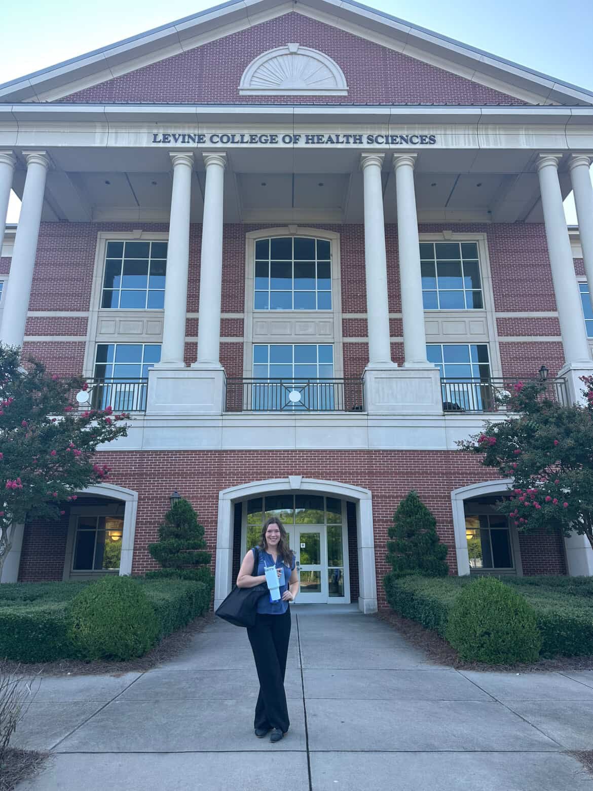 A woman smiling and standing outside a red brick building labeled Levine College of Health Sciences, holding a folder or notebook, with bushes and trees around the entrance.