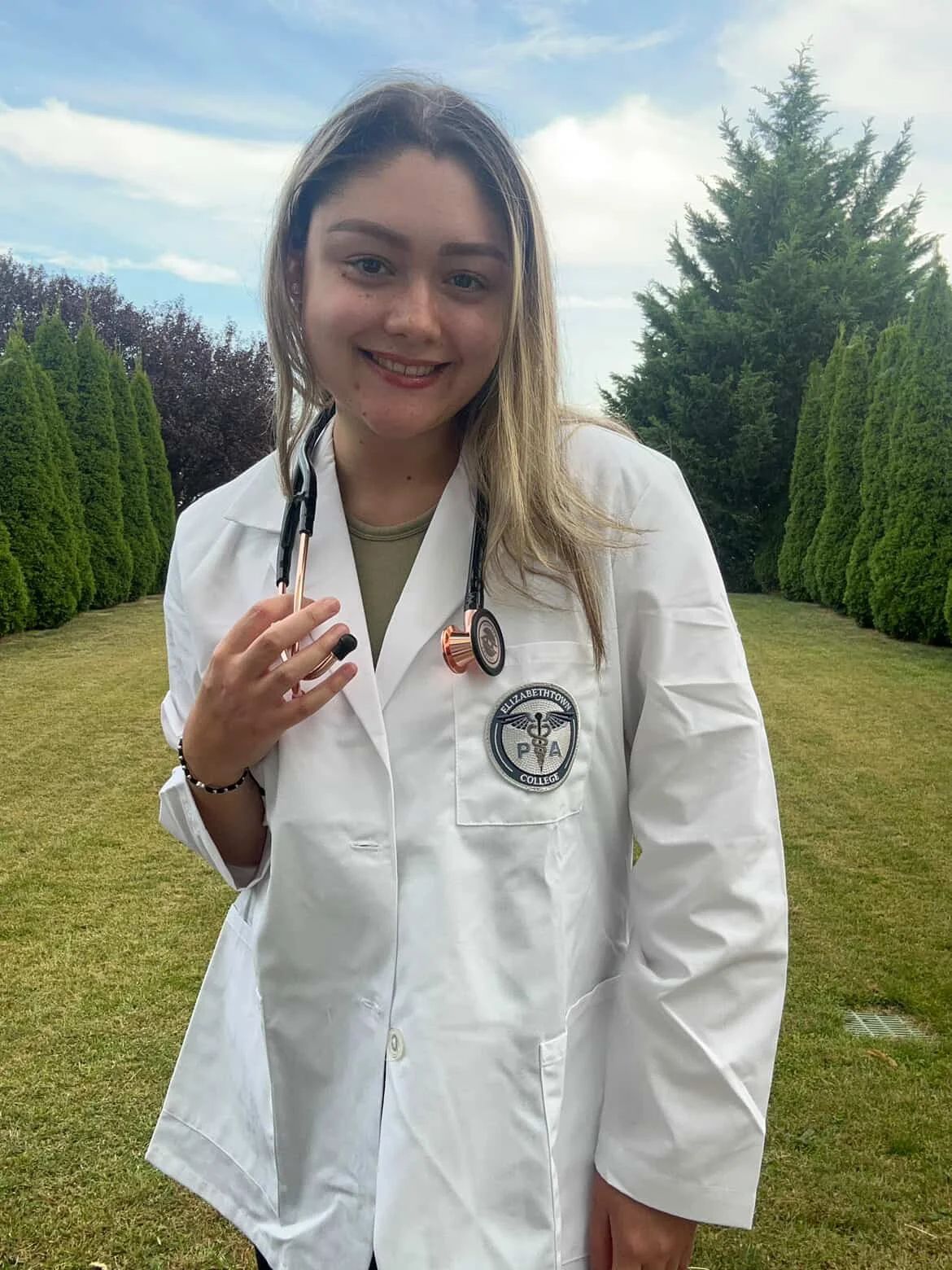 Young woman in white medical coat with stethoscope outdoors, green trees and cloudy sky in background.