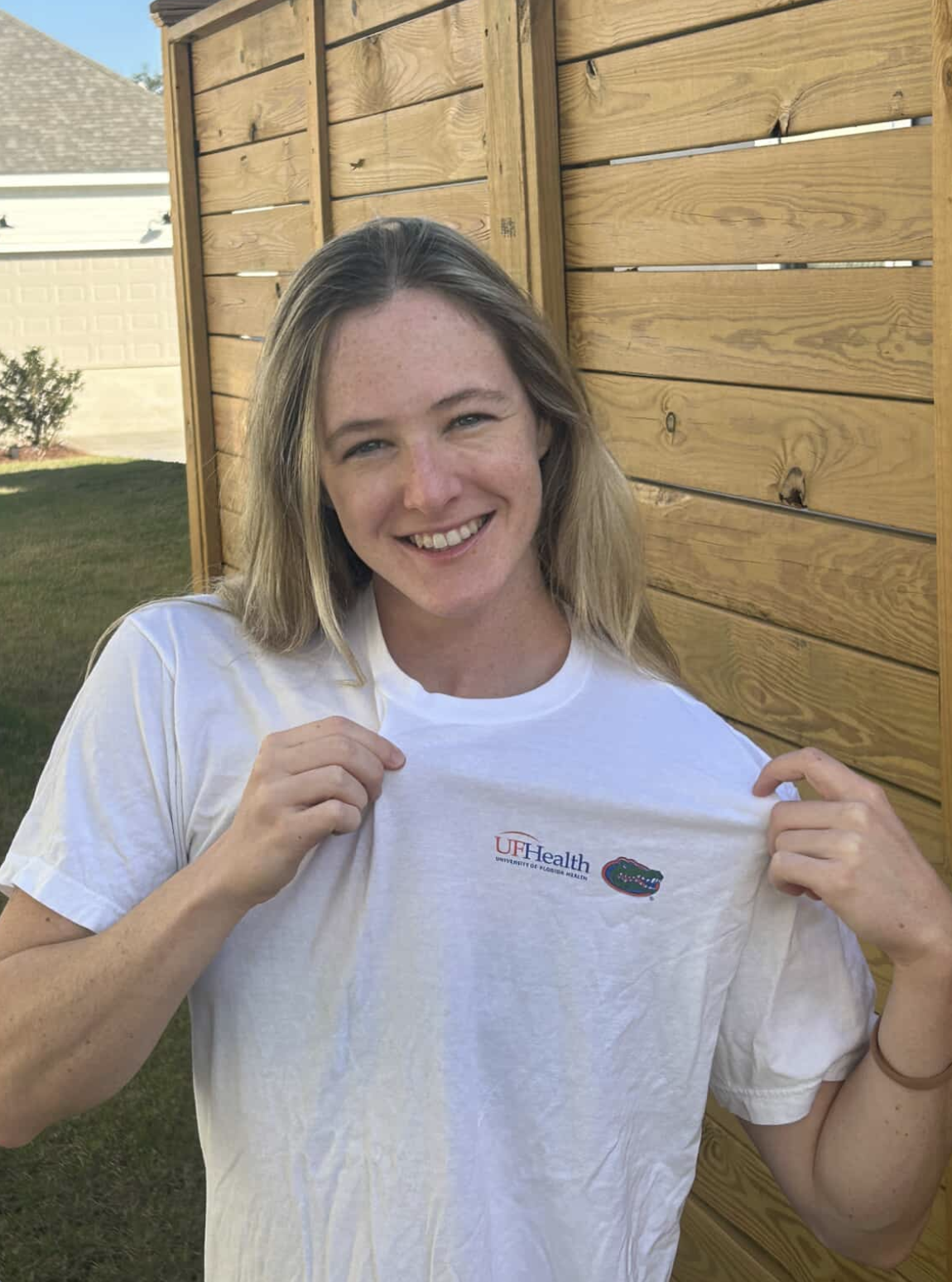 A woman smiling and showing a white T-shirt with UF Health University of Florida Health logo and alligator emblem in front of a wooden fence.