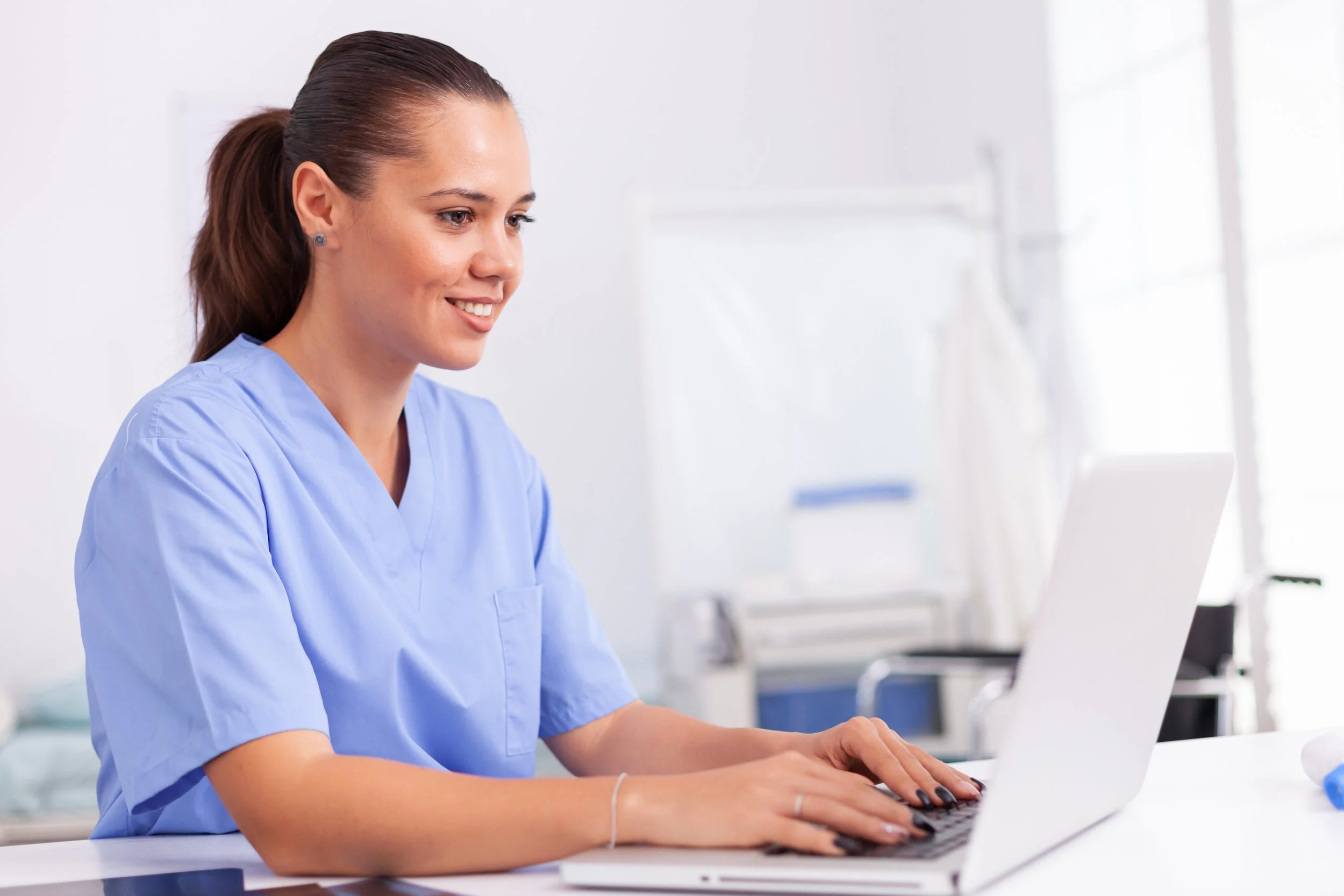 A female nurse in blue scrubs working on a laptop in a clinical setting.