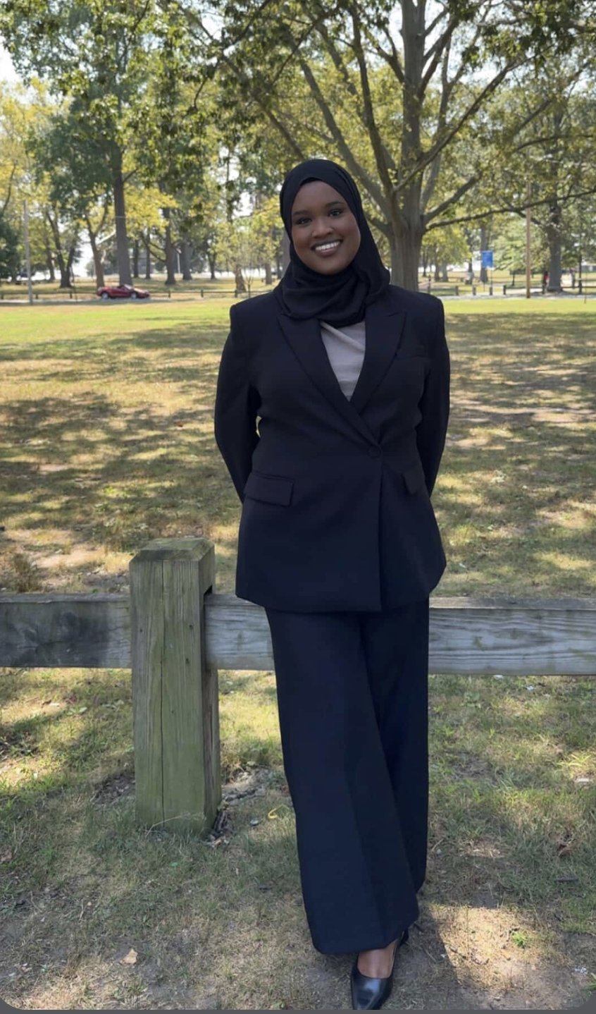 A woman in a black suit and hijab standing outdoors in a park with trees and grass.