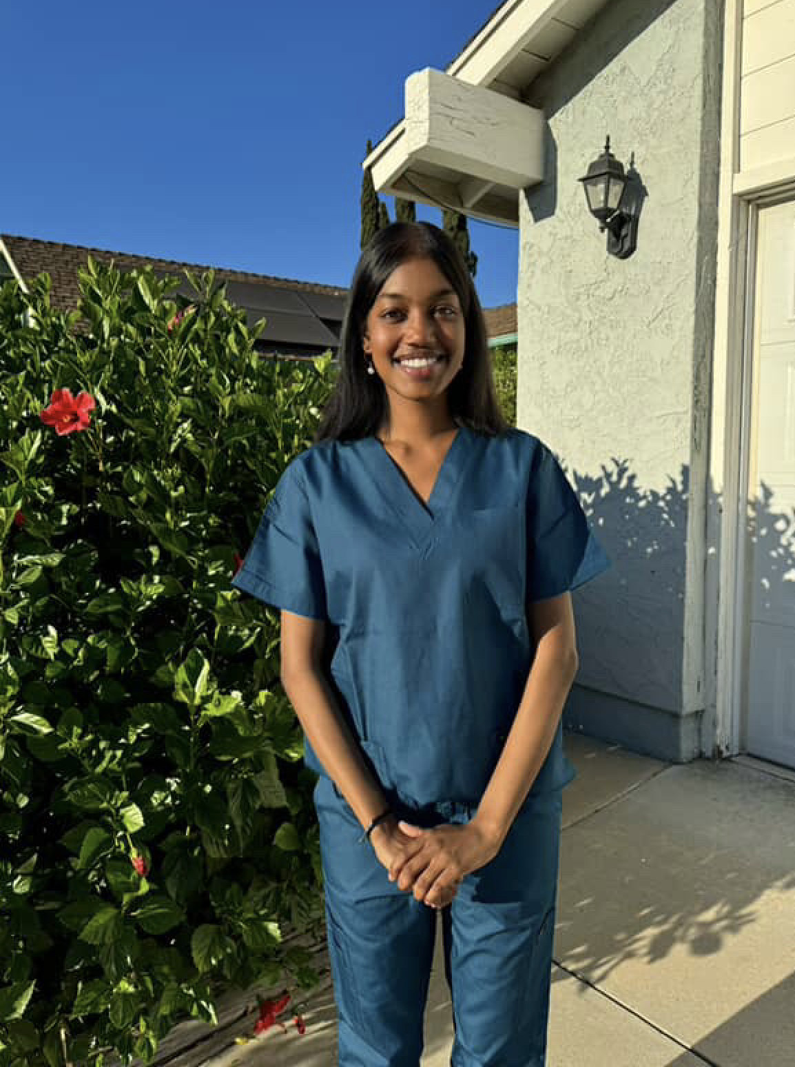 A young woman wearing blue medical scrubs standing outdoors on a sidewalk next to a green bush with pink flowers, smiling at the camera, with a house wall and a light fixture in the background.