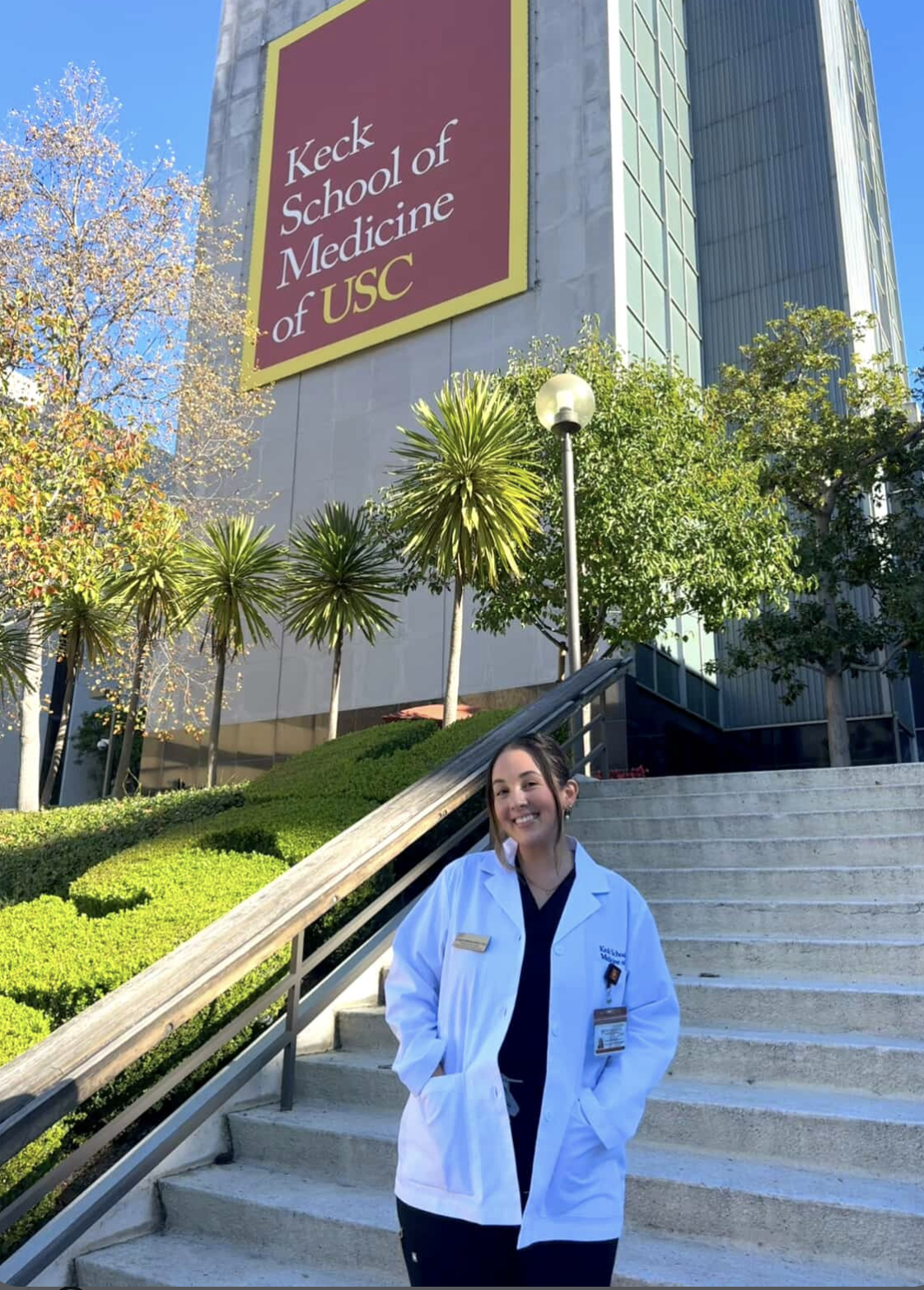 A young woman in a white medical coat standing on stairs outside the Keck School of Medicine of USC, with greenery and a large building with a sign reading "Keck School of Medicine of USC" in the background.