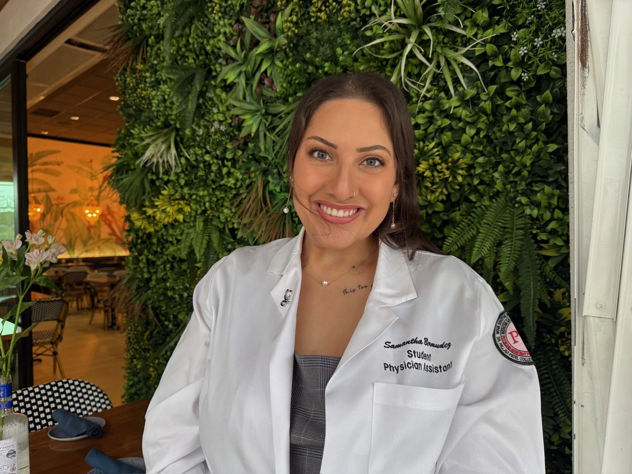 A young woman with long dark hair, wearing a white medical coat, smiling in front of a green plant wall inside a restaurant or cafe.