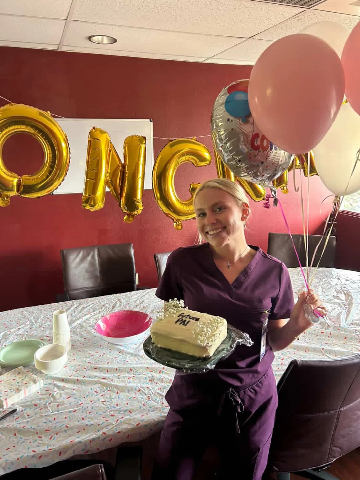 A woman in scrubs holding balloons and a cake at a birthday celebration for a father, with gold letter balloons spelling out 'PAPA' in the background.