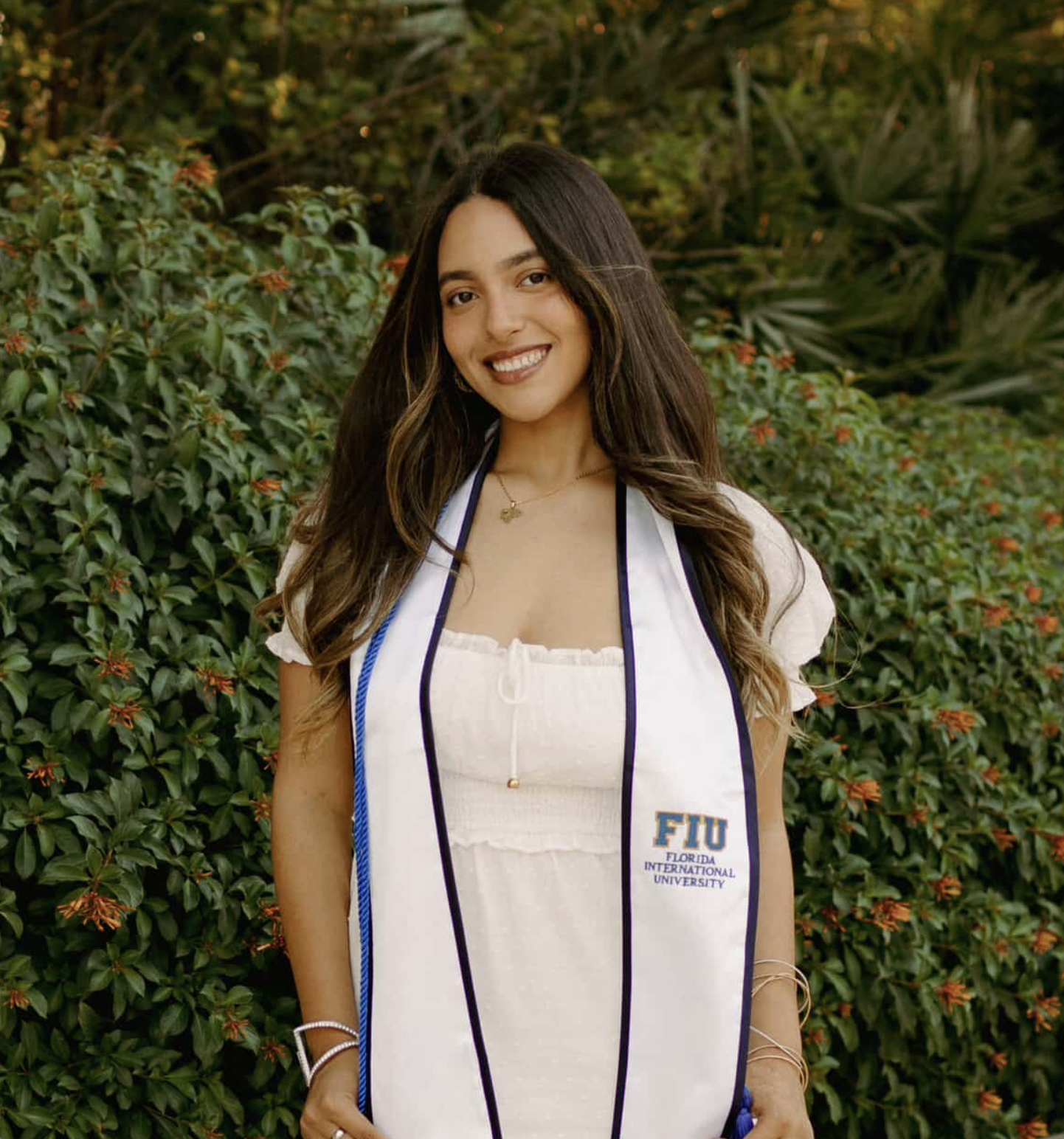 Young woman outdoors wearing a graduation stole from Florida International University, smiling with a bush and trees in the background.