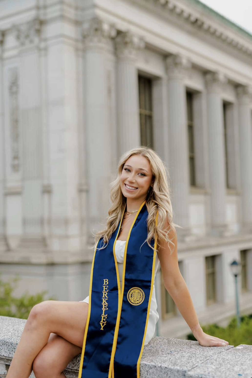 A young woman with long blonde hair, wearing a white dress and a blue and gold graduation stole that says 'Berkeley,' sitting outdoors on a stone ledge in front of a historic building with columns, smiling at the camera.