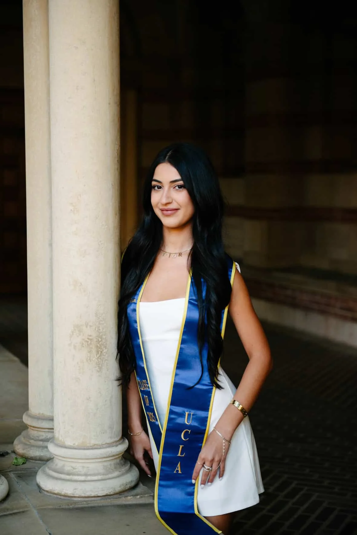 Young woman with long dark hair, wearing a white dress and a blue sash with gold trim that reads 'UCLA', standing outdoors next to a stone column.