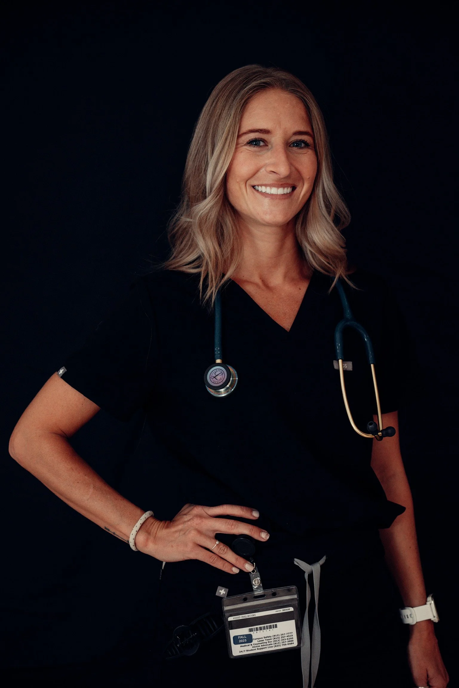 A smiling female healthcare professional wearing black scrubs with a stethoscope around her neck, standing against a dark background.