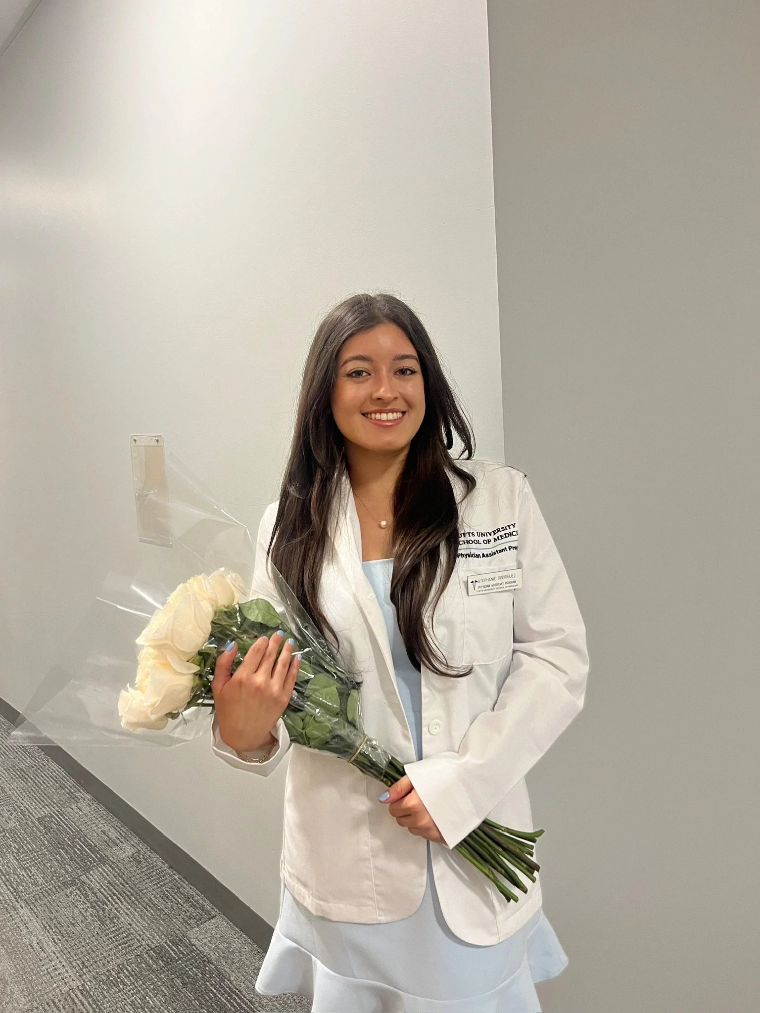 A young woman in a medical white coat holding a bouquet of white roses, standing in front of a plain wall and smiling at the camera.