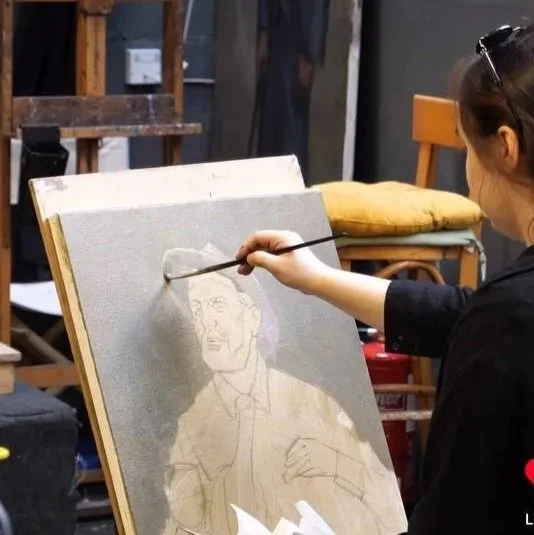 A person with sunglasses on their head painting a sketch of a person on a canvas with a paintbrush, in an art studio.