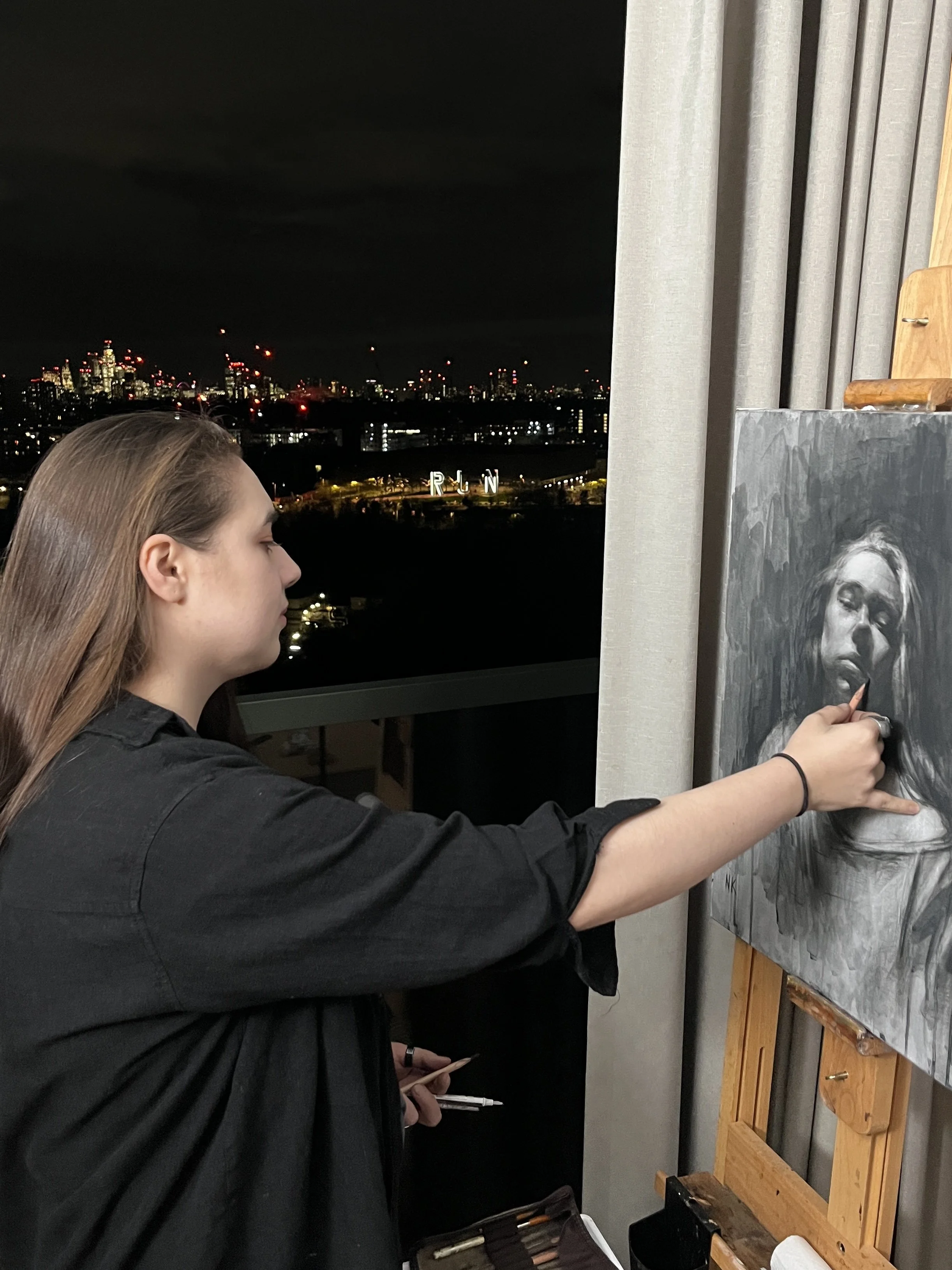 A woman painting a black and white portrait of a woman, in an indoor setting at night, with a city skyline visible through a window in the background.