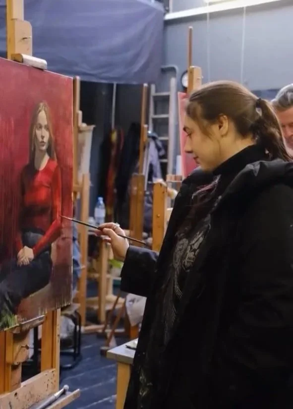 A woman with dark hair tied back is painting a portrait of a woman in a red shirt on a canvas in an art studio.