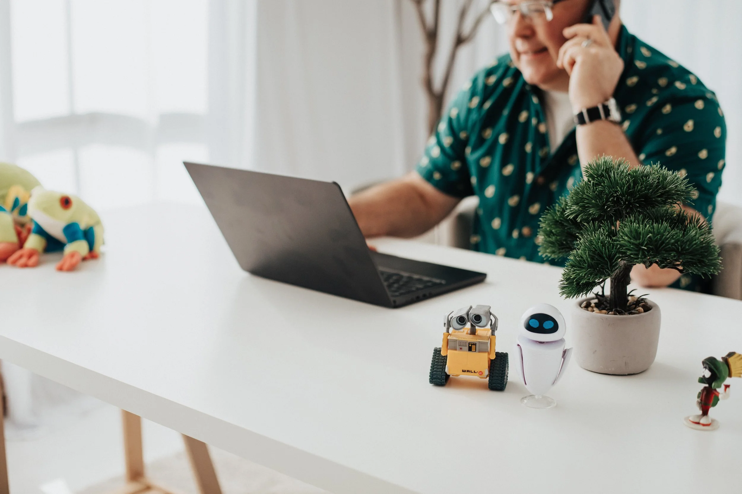 An elderly woman sits at a white table talking on a phone while looking at her laptop. The table has a small potted pine tree, a small robot, a white robot with blue eyes, a frog plush toy, and a small Santa figurine.