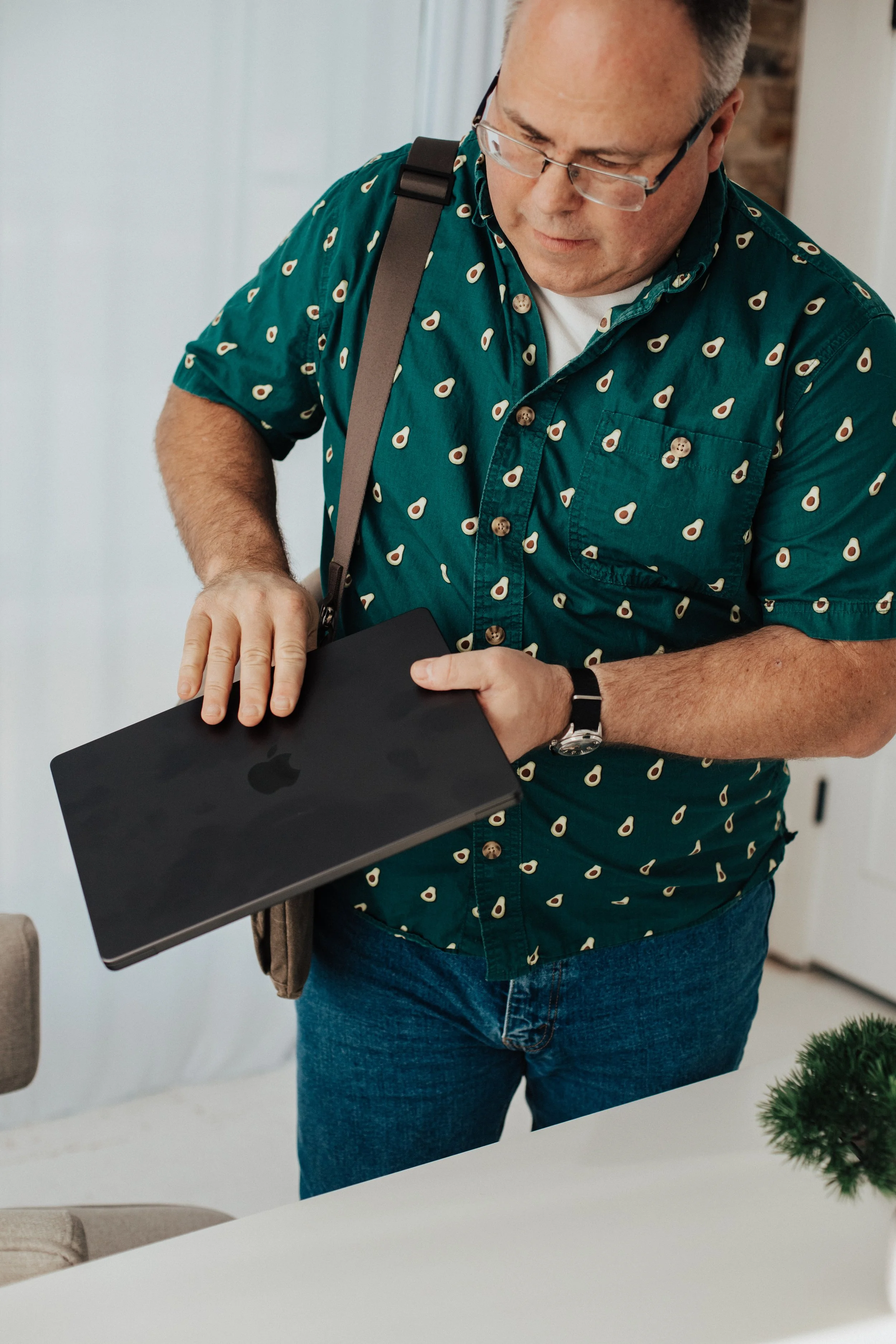 A man wearing a teal shirt with a pattern of avocados is holding a closed black MacBook laptop.